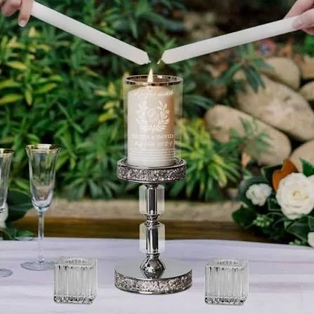 A lit white candle in a glass holder on a decorative silver stand, with two unlit white candles being held above it, outdoors with greenery and flowers in the background.