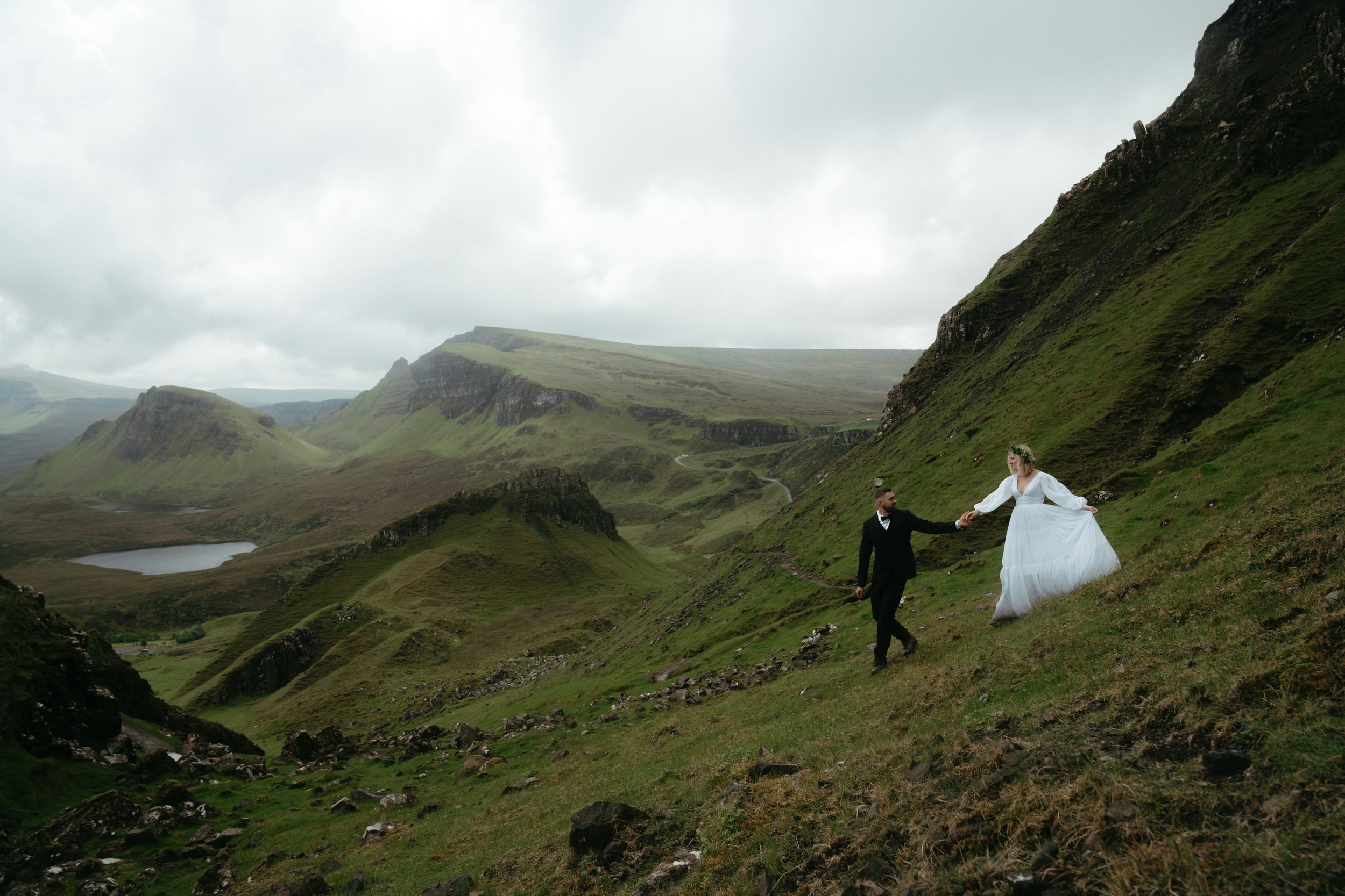 A bride and groom holding hands while dancing outdoors in a mountainous landscape with green rolling hills and cloudy sky.