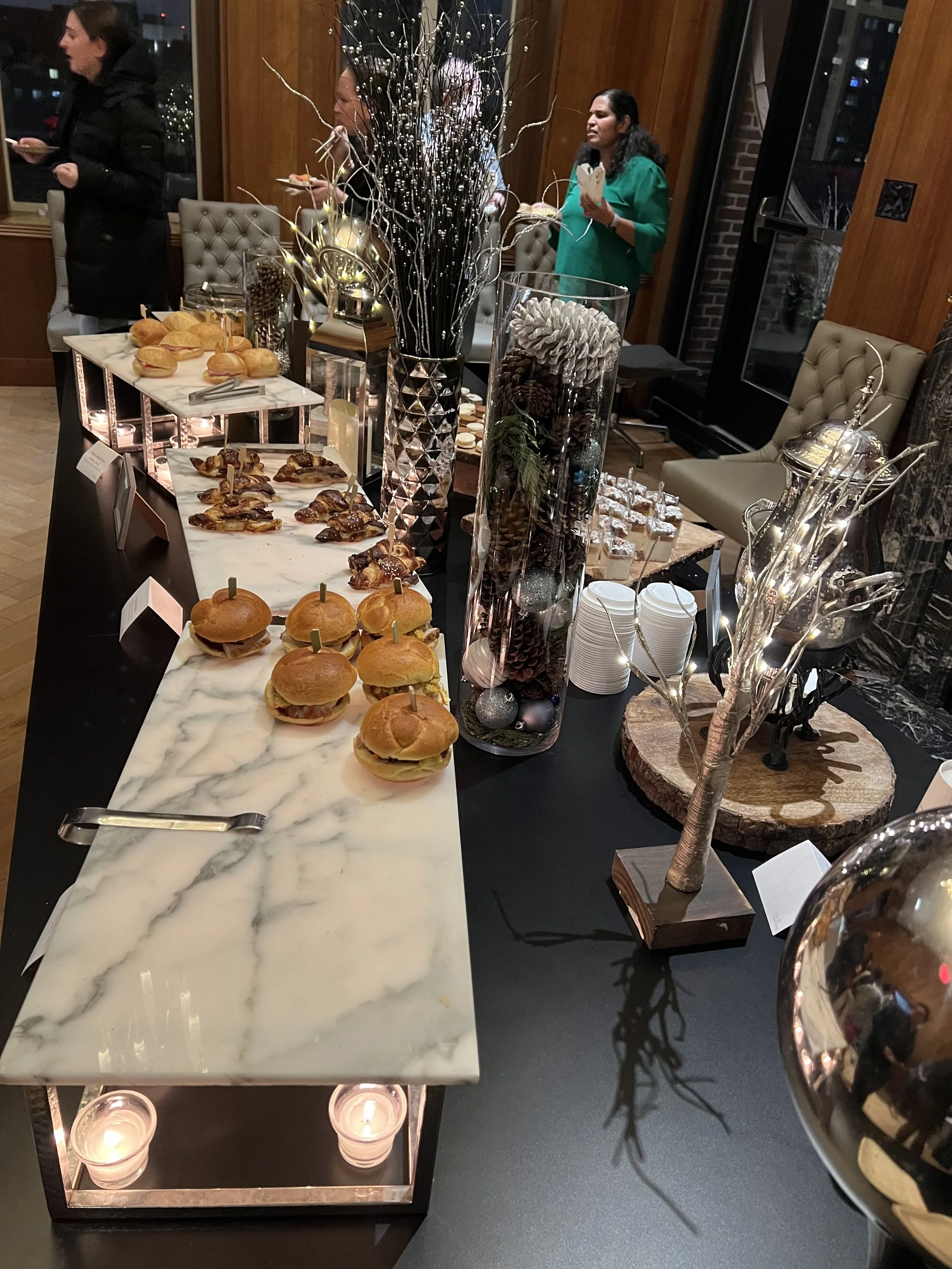 A winter holiday party spread with buffet table of mini burgers, chicken wings, and desserts, decorated with candles, vases filled with pinecones and festive branches