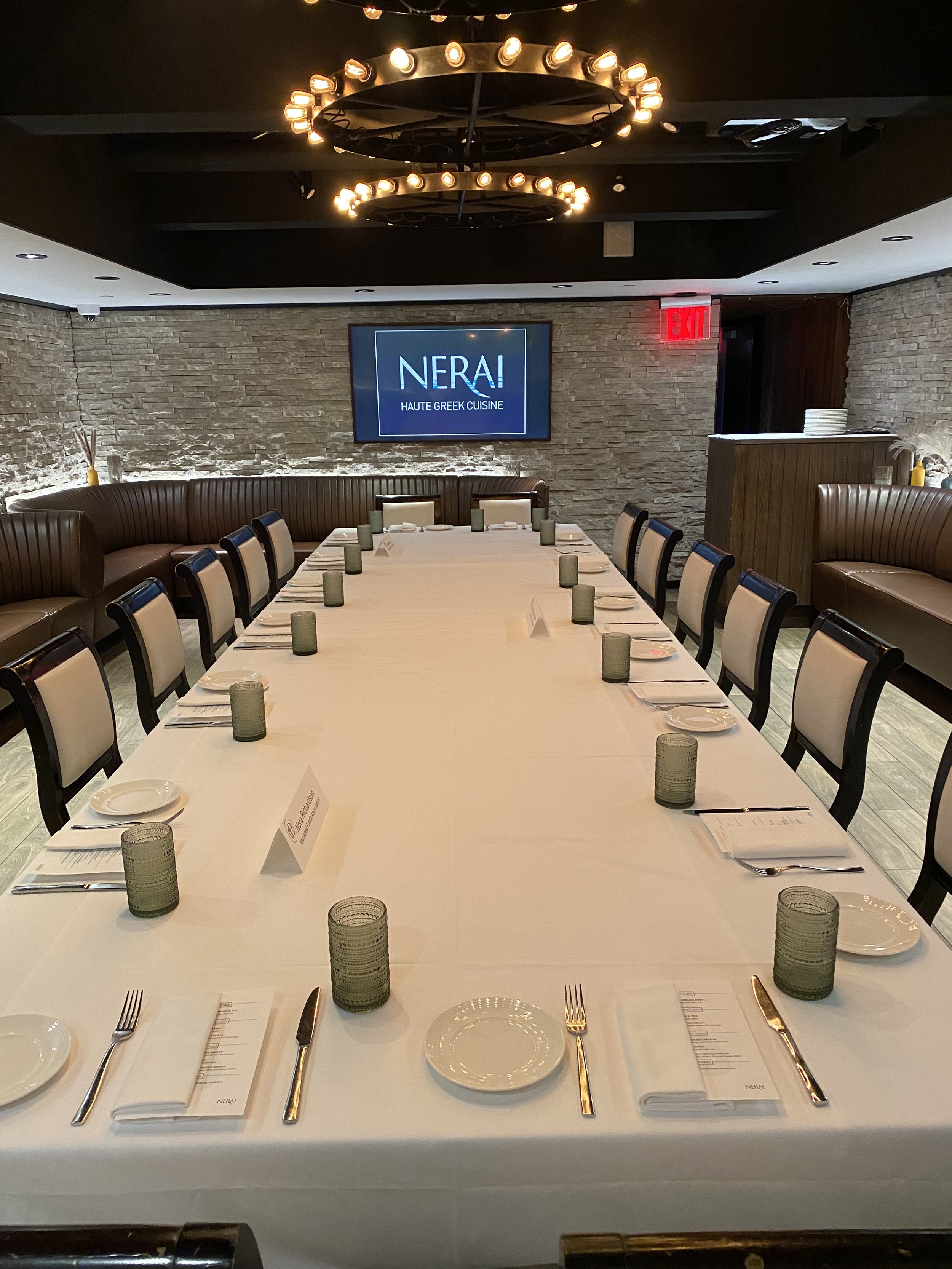 A formal dining table setup at Nerai restaurant with white tablecloth, plates, silverware, napkins, green glasses, and menus, in a restaurant with stone walls and modern lighting fixtures.
