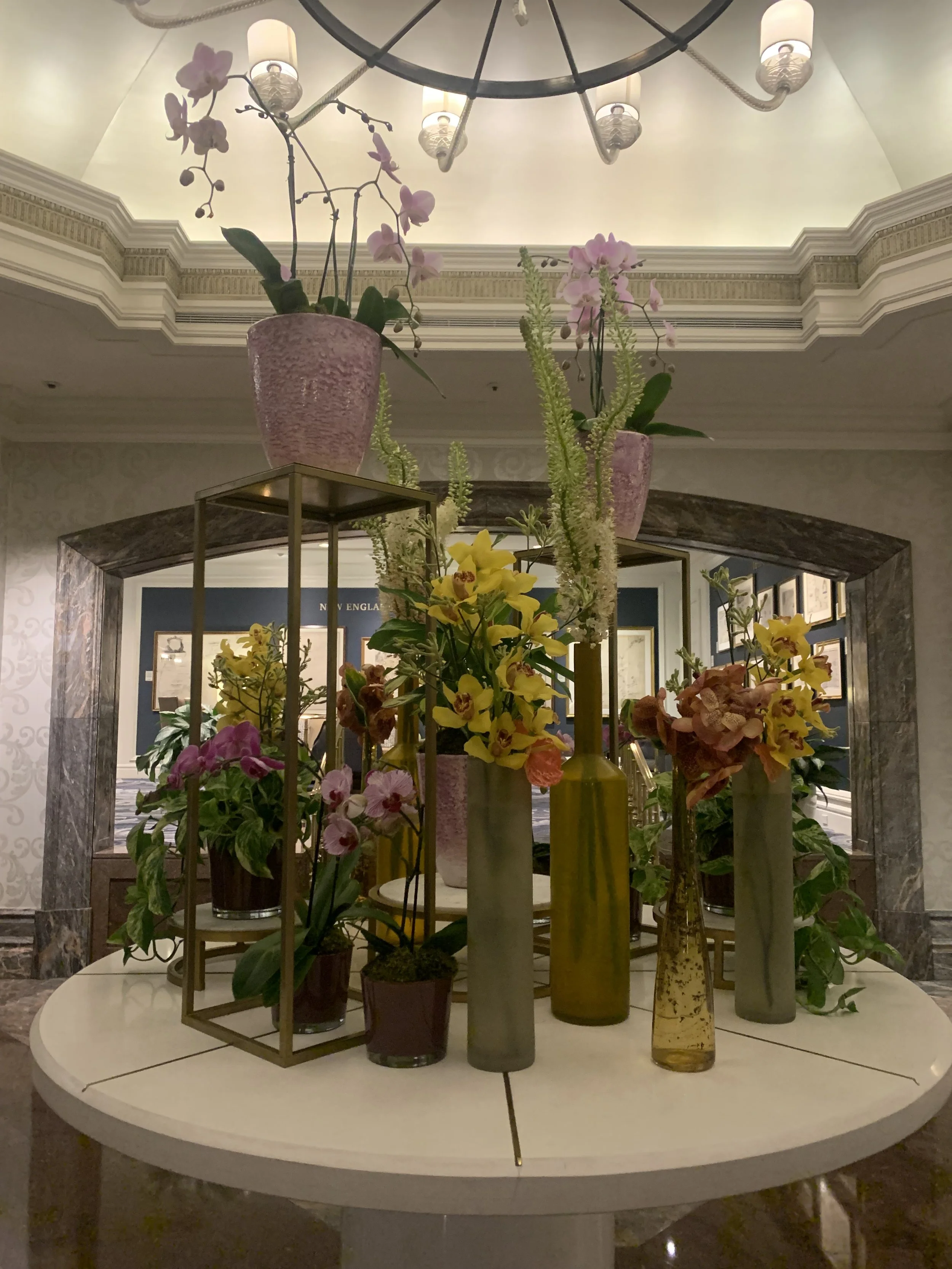 A decorative display of assorted potted and vase flowers, including orchids and other blooms, arranged on a stand in front of an archway and a lobby wall.