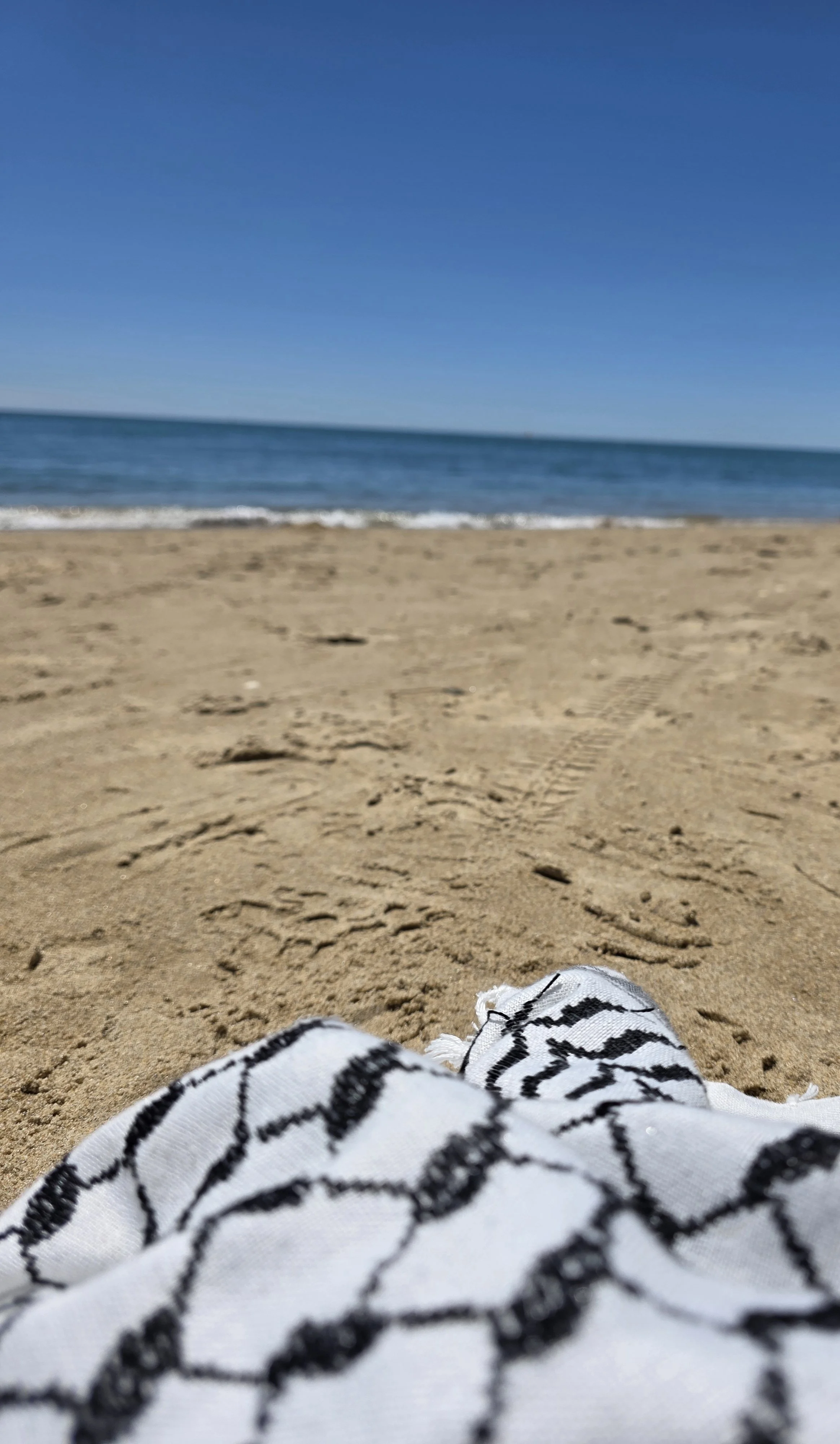 Vista de una playa con arena y mar, vista desde una persona acostada en la arena con sandalias blancas y rosas, en un día soleado con cielo despejado. Kufiya. Pañuelo Palestino.