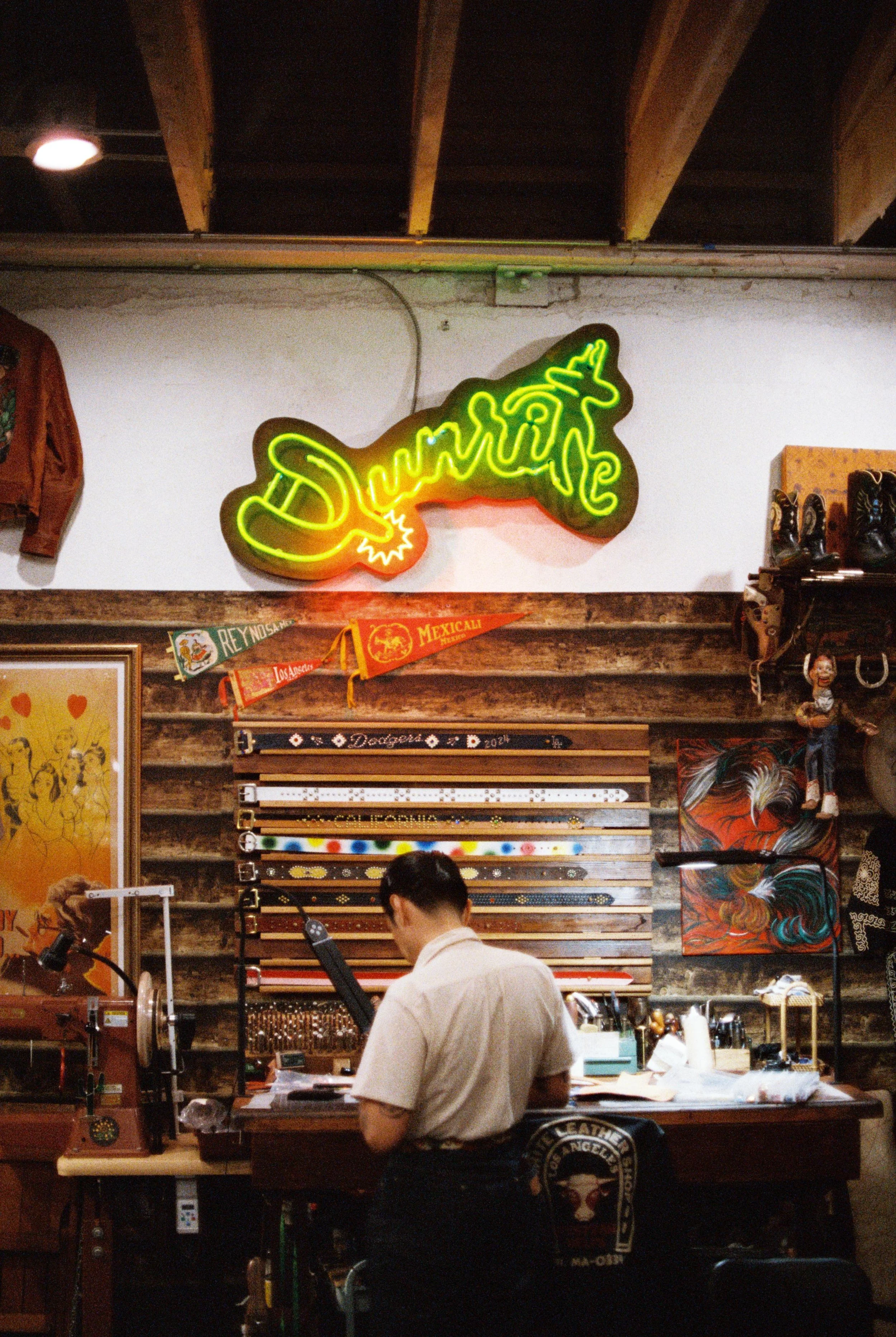Neon sign with the word 'Drive' on a white wall, surrounded by various items in a store, including artwork, belts, and clothing.