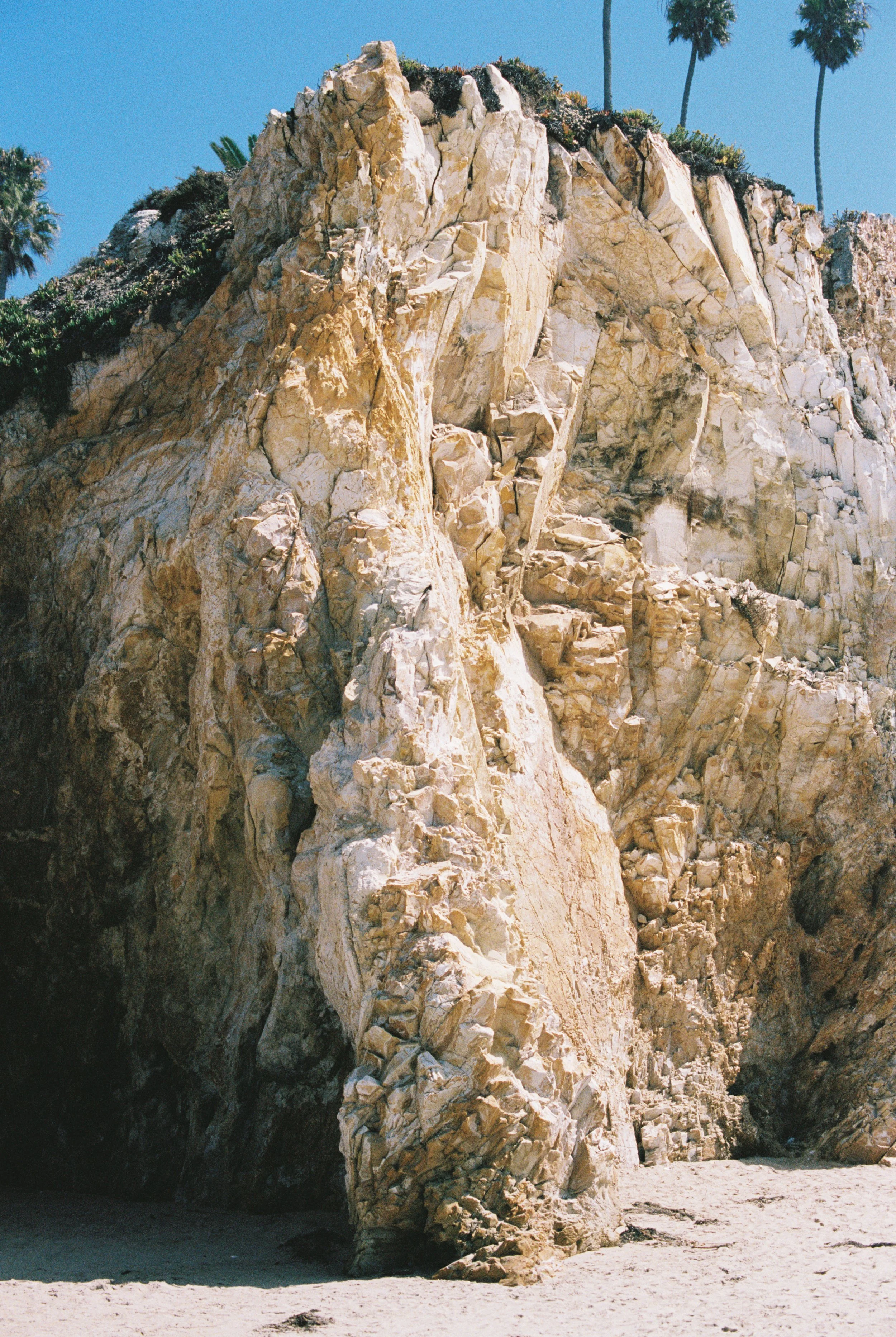 Large beige rock formation surrounded by palm trees and a sandy beach under a clear blue sky.
