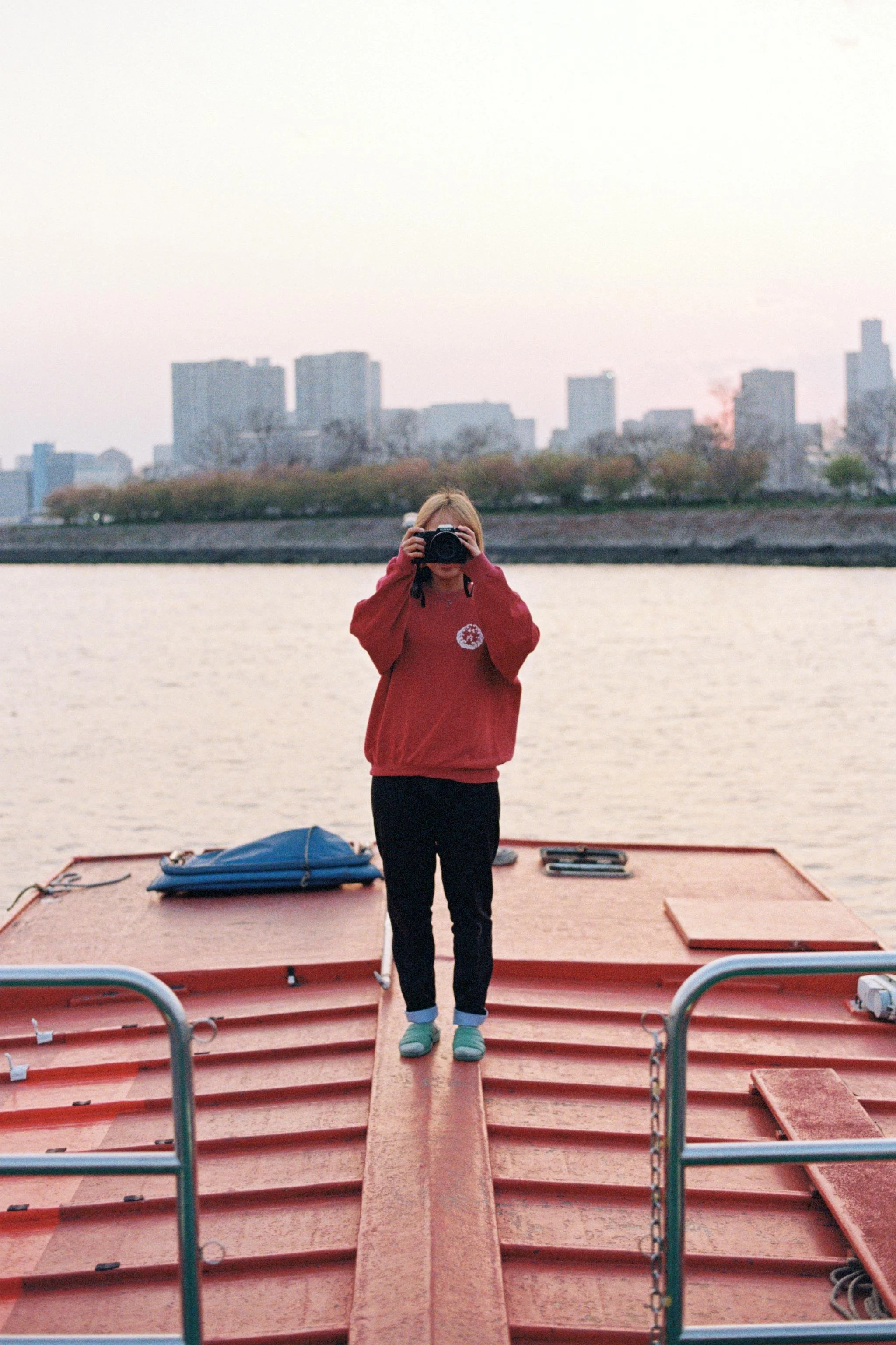 Person standing on a boat taking a photo with a camera, calm water, city skyline in the background during dusk or dawn.