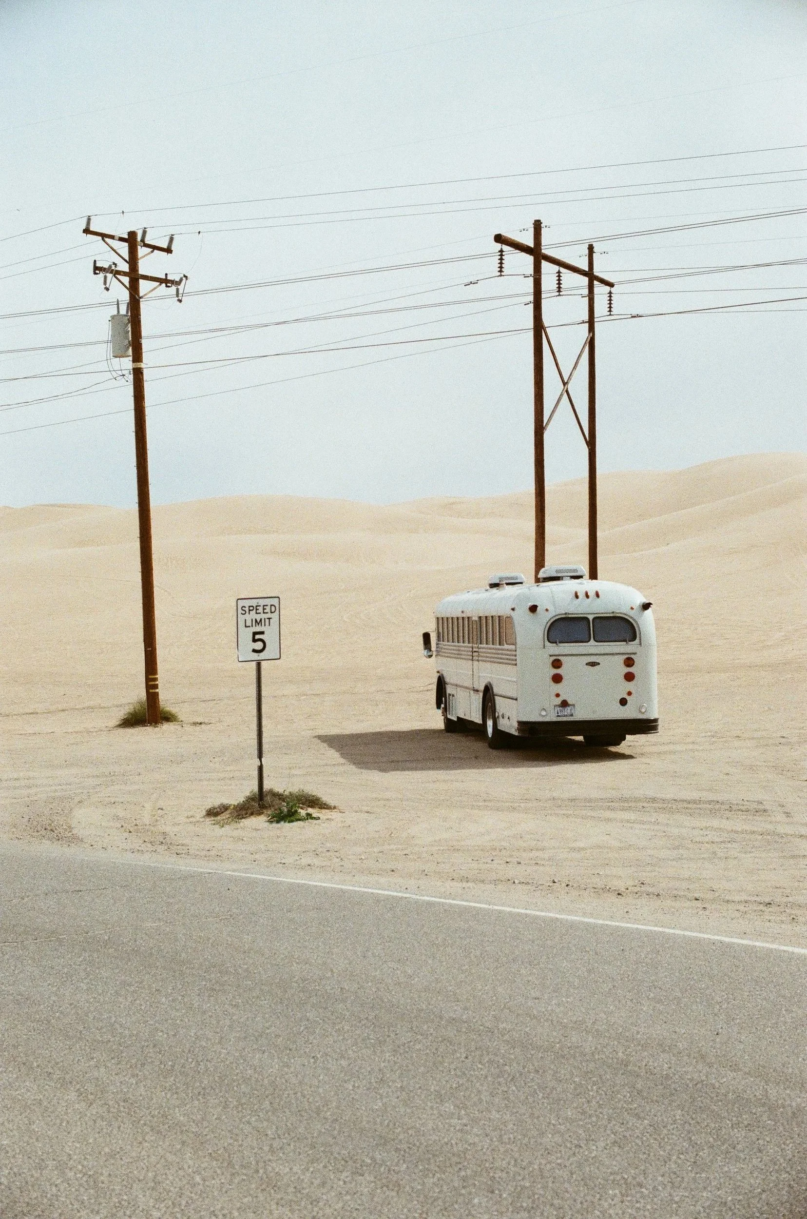 A desert scene with sand dunes and power lines, a speed limit sign of 5 mph, and an old bus parked on the side of the road.