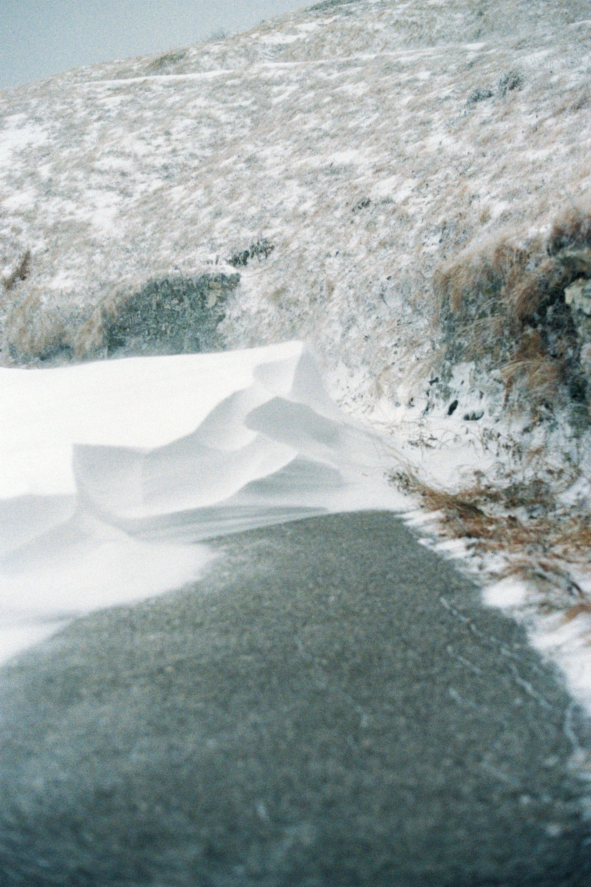 Snow-covered ground with a rocky hillside in the background and a shadow of the person taking the photo in the foreground.