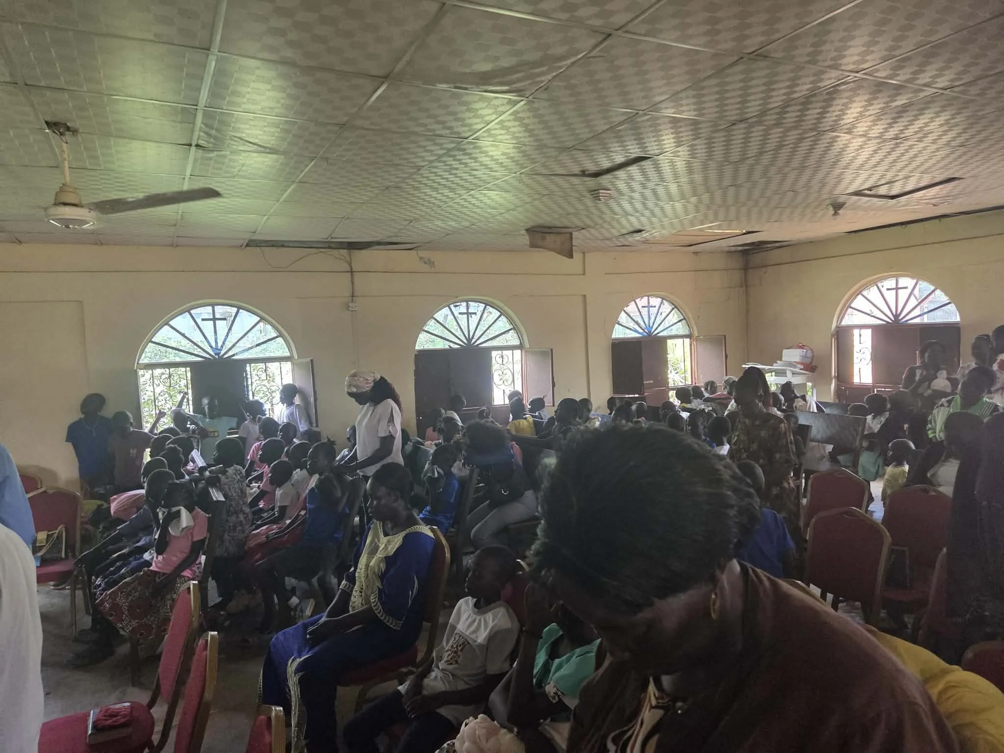 A crowded indoor gathering with many children and some adults, seated and standing, inside a room with arched windows and a patterned ceiling, during daytime.
