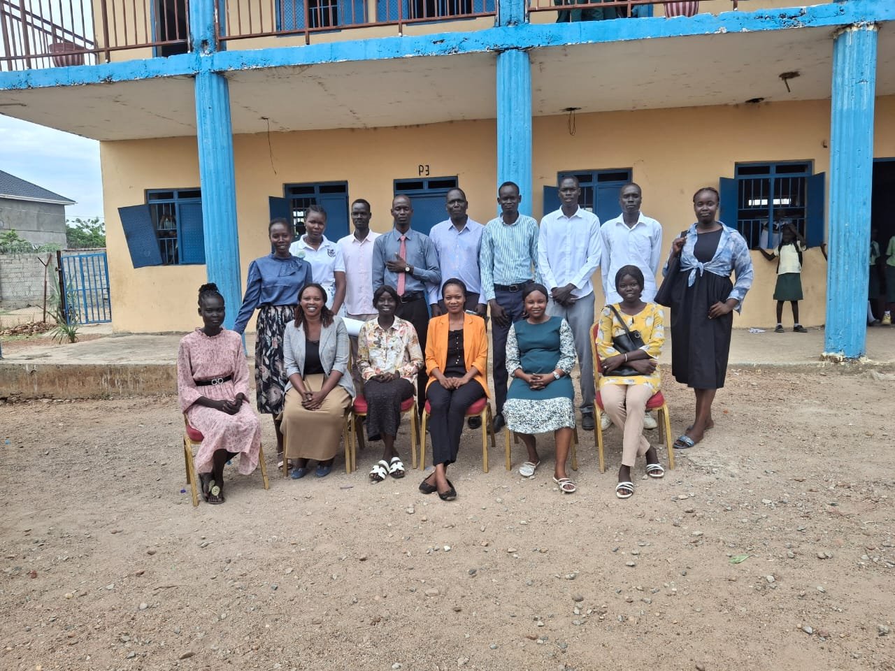 Group of fifteen people, mostly women, posing for a photo outside a yellow building with blue doors and railings, in a rural area.