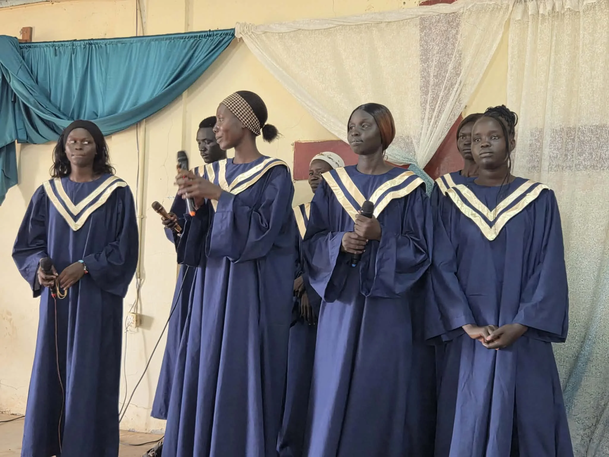 Several women dressed in dark blue robes with cream and gold trim are standing in a line, some holding microphones. They are indoors, against a plain wall with some drapery, and appear to be participating in a formal event.