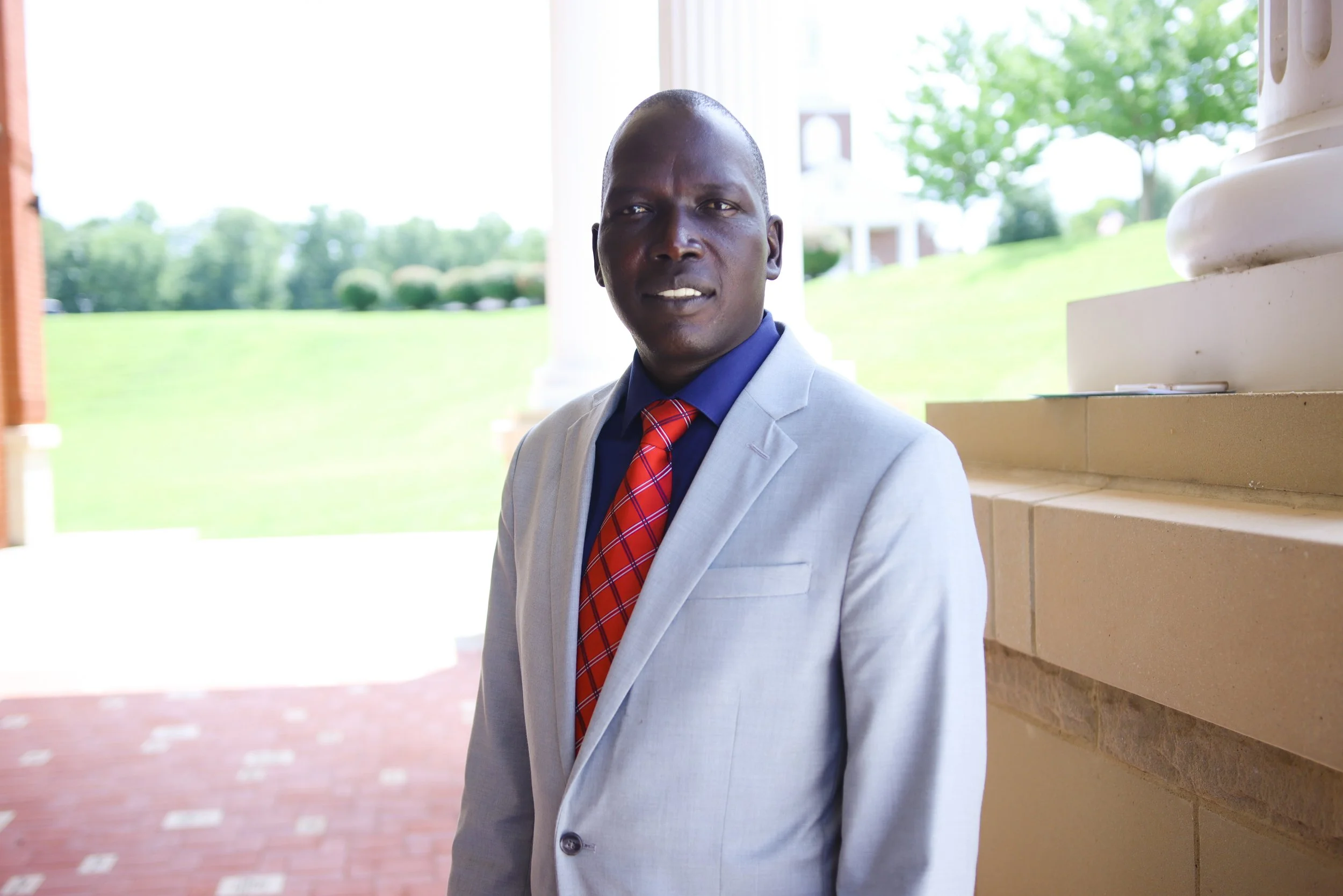 A man wearing a light gray suit, blue shirt, and red plaid tie standing outdoors on a porch with greenery in the background.