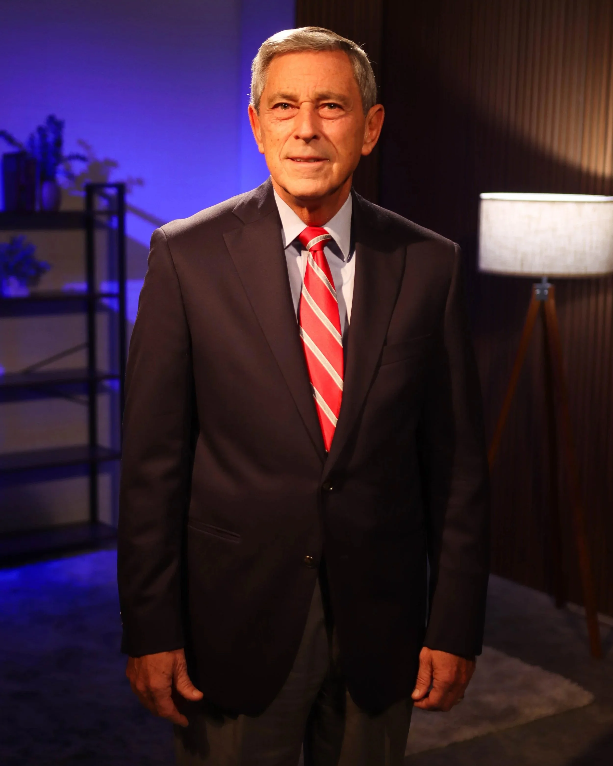 Older man wearing a dark suit, white shirt, and red striped tie, standing indoors with a lamp and a bookshelf in the background.