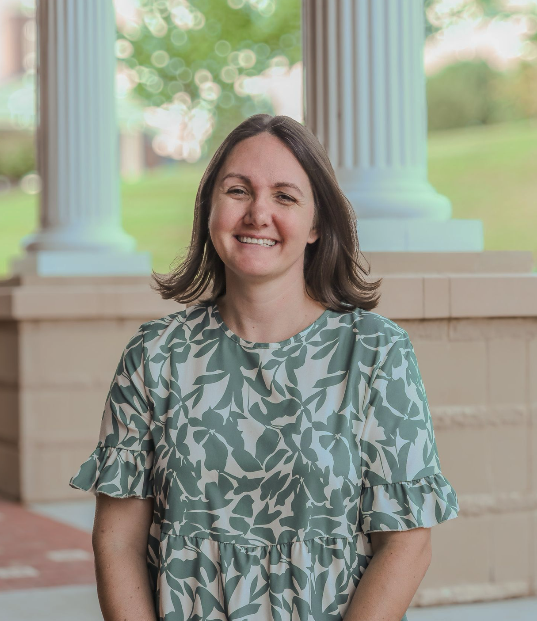 Woman with shoulder-length brown hair, smiling, standing in front of classical columns with a green outdoor background.