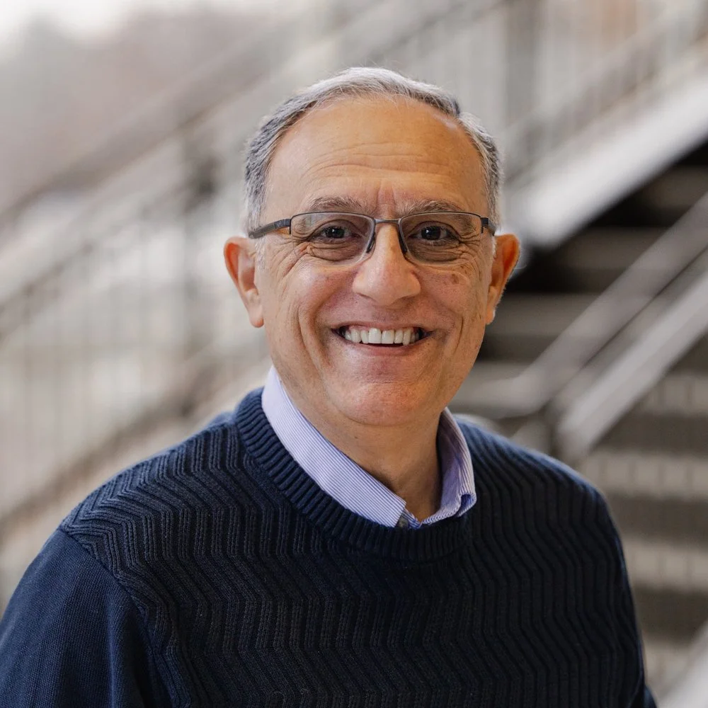 A smiling middle-aged man with gray hair, wearing glasses, a purple collared shirt, and a navy blue sweater, standing on a staircase in a building with a metal railing.