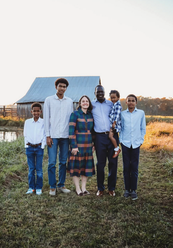 A family of six standing outdoors in front of a blue barn, with grass and a pond nearby, during sunset.
