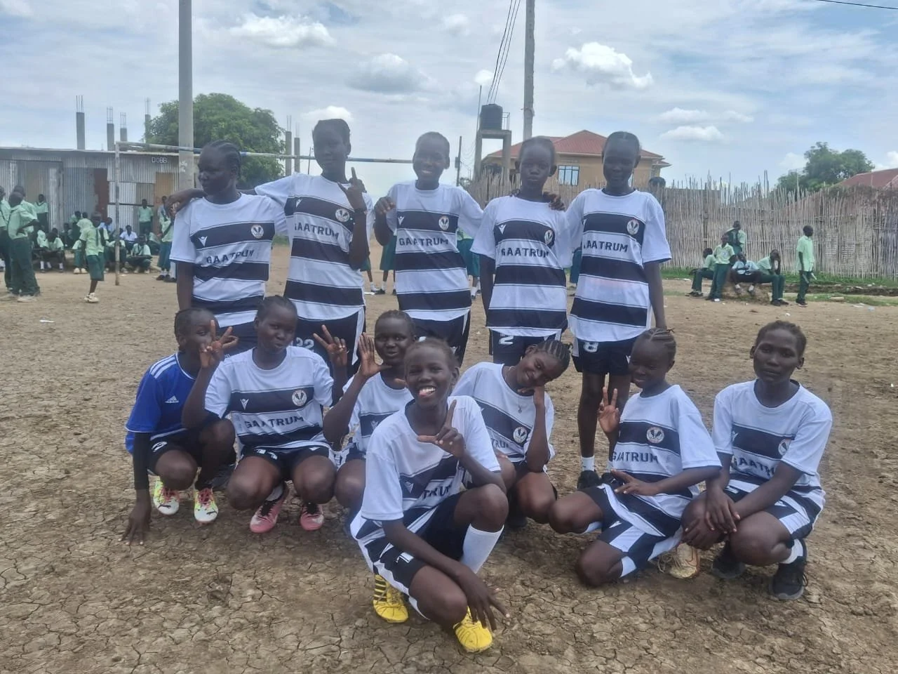 Group of children in soccer uniforms posing on a dirt field, some making peace signs, with other children and a building in the background.