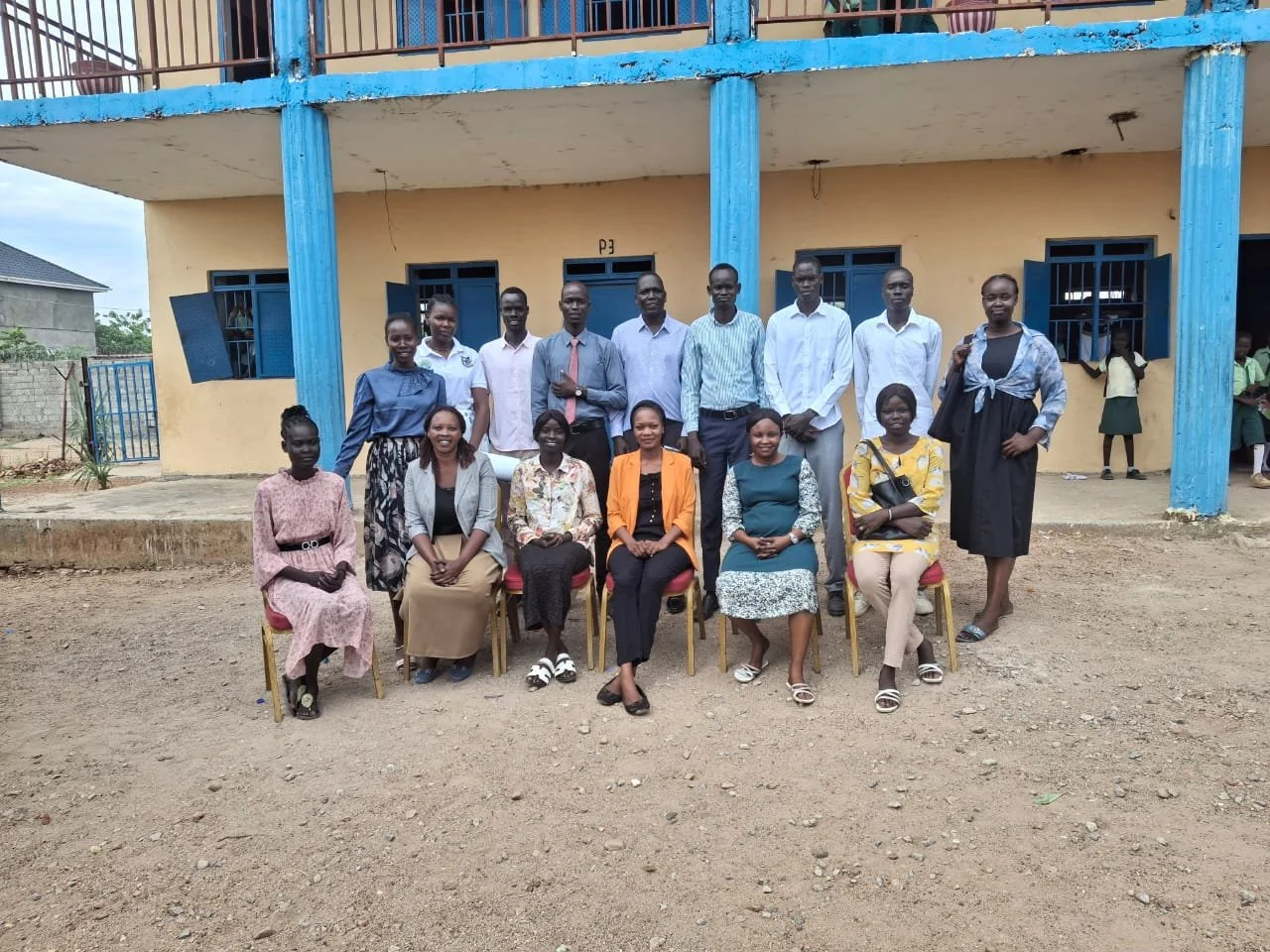 A group of 15 people, including men and women, posing for a photo outside a school building. The building has a yellow wall with blue window frames and pillars. The ground is unpaved.