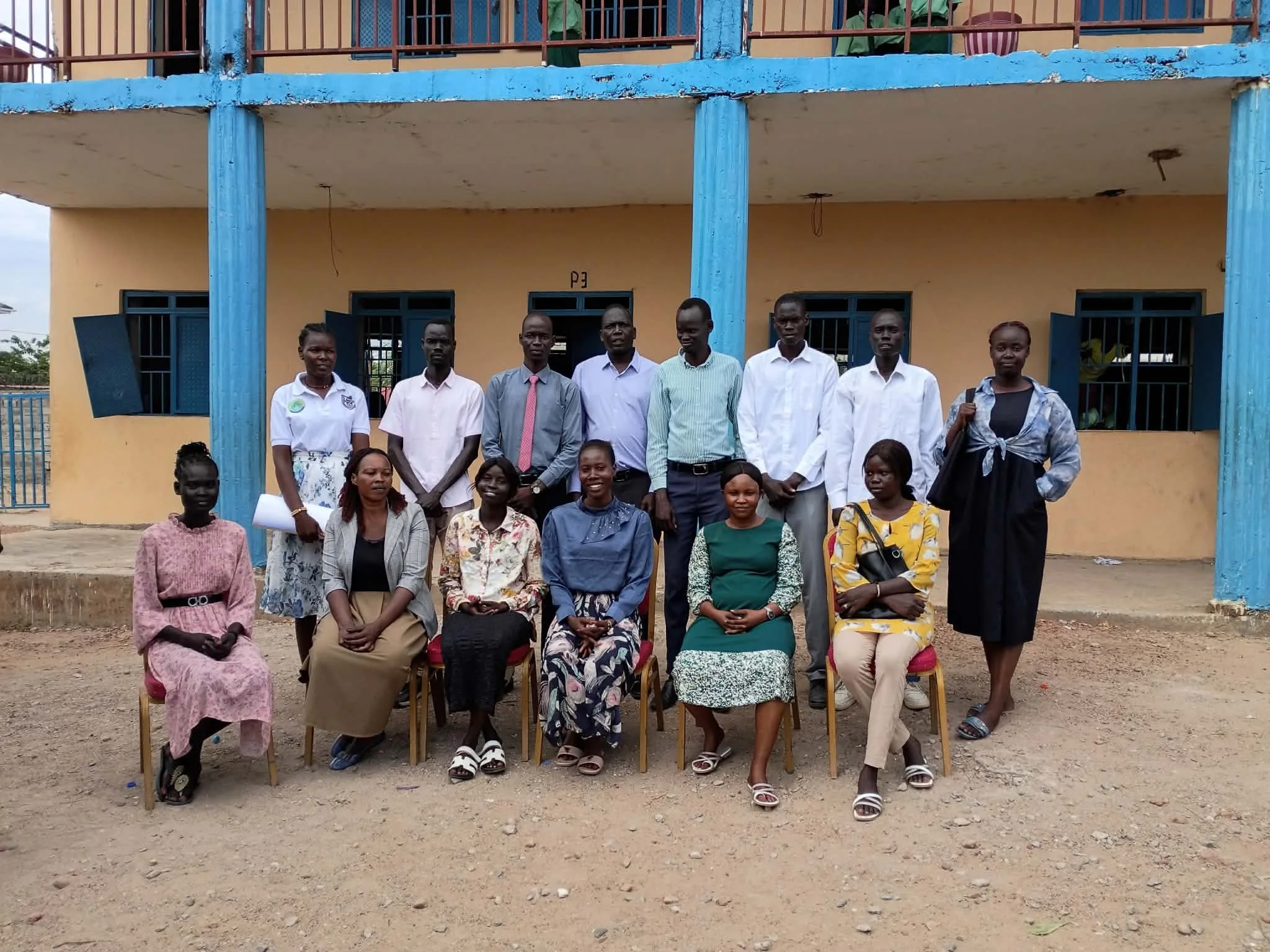 A group of adults and children posing for a photo outside a yellow building with blue columns and railings. Some people are standing, and some are seated on chairs in front of the building.