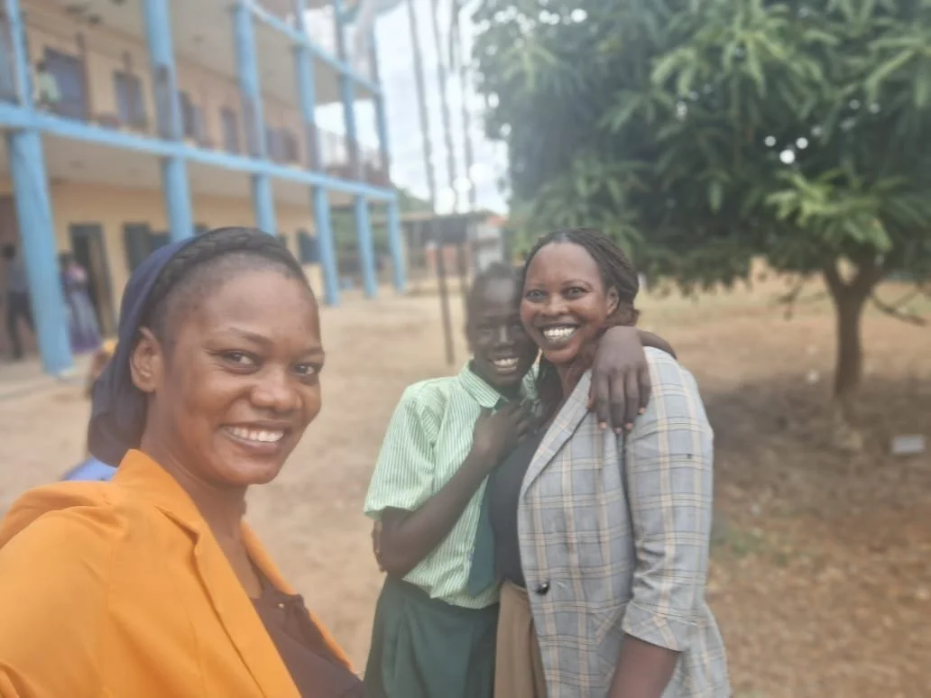 Three smiling women and a boy standing outdoors near a school building and trees in a dry area.