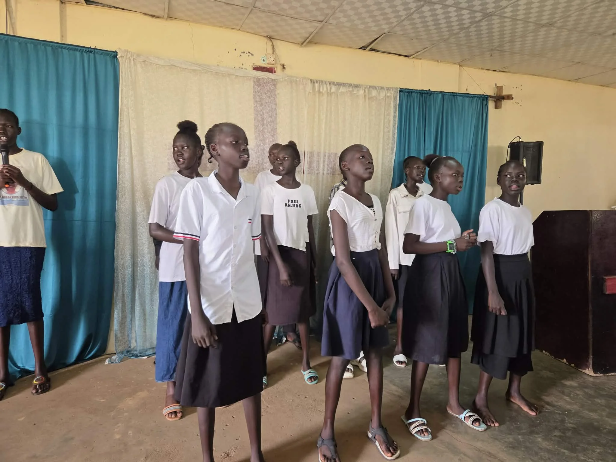 A group of young girls standing on stage in a school setting, facing forward, with some singing or reciting. The girls are wearing uniforms, and the stage is decorated with blue and beige curtains. A speaker and a cross are visible in the background.