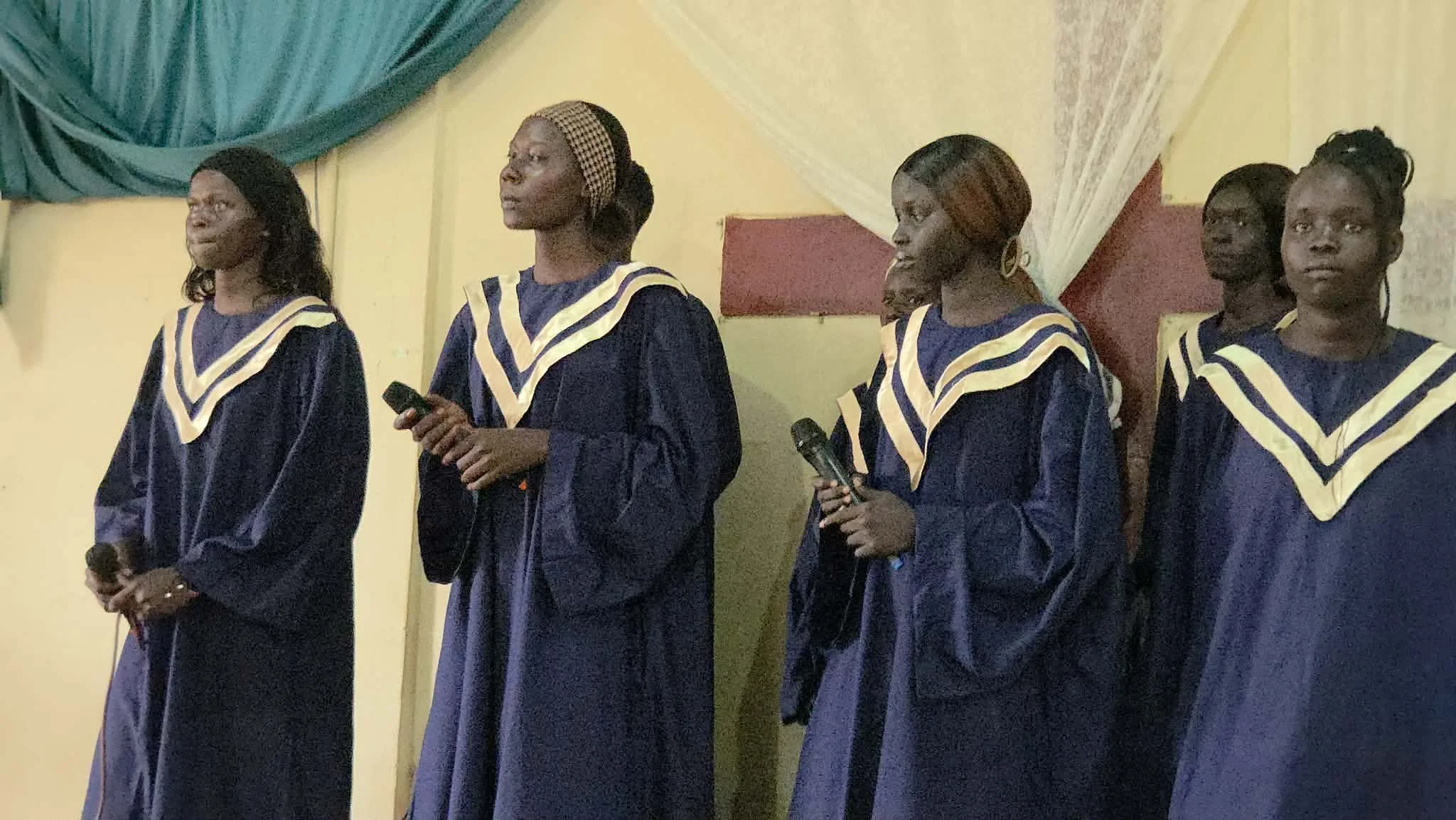 Four women in matching graduation gowns and stoles standing in a line at a graduation ceremony, holding microphones.
