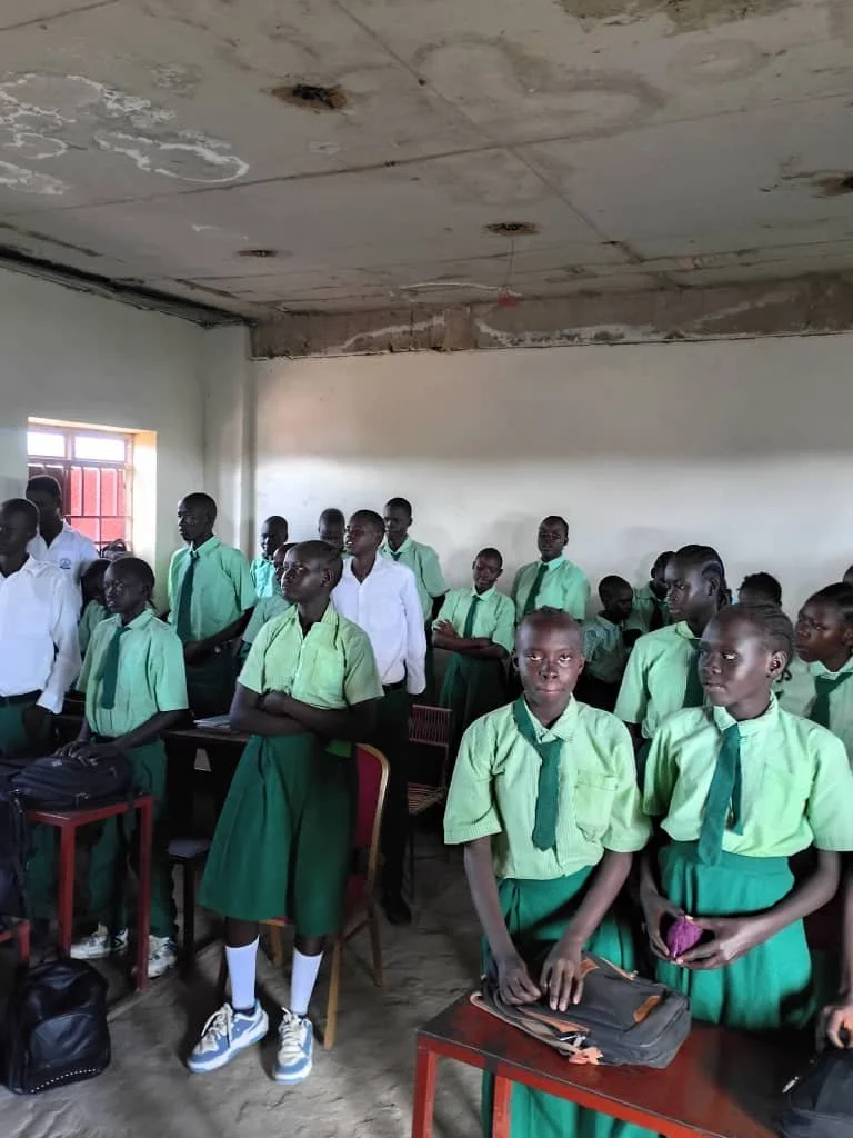 Group of students in green uniforms and ties standing inside a classroom with a partially finished ceiling and a window with red bars.