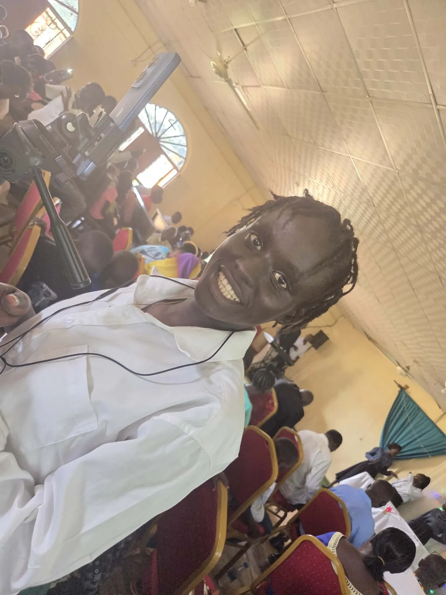 Young man smiling in white shirt standing in a crowded event hall with seated audience and a camera on a tripod in the foreground.