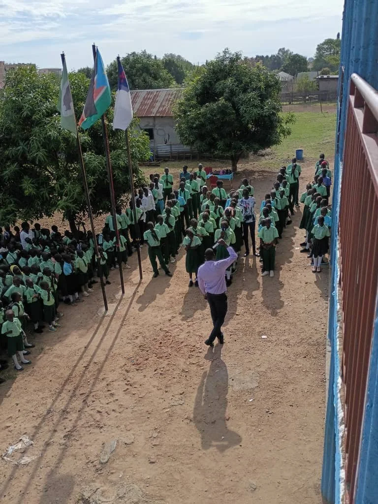 School assembly with students in green uniforms, students standing in lines outside, flags on poles, teacher or administrator in front, outdoor schoolyard with trees and a building in the background.