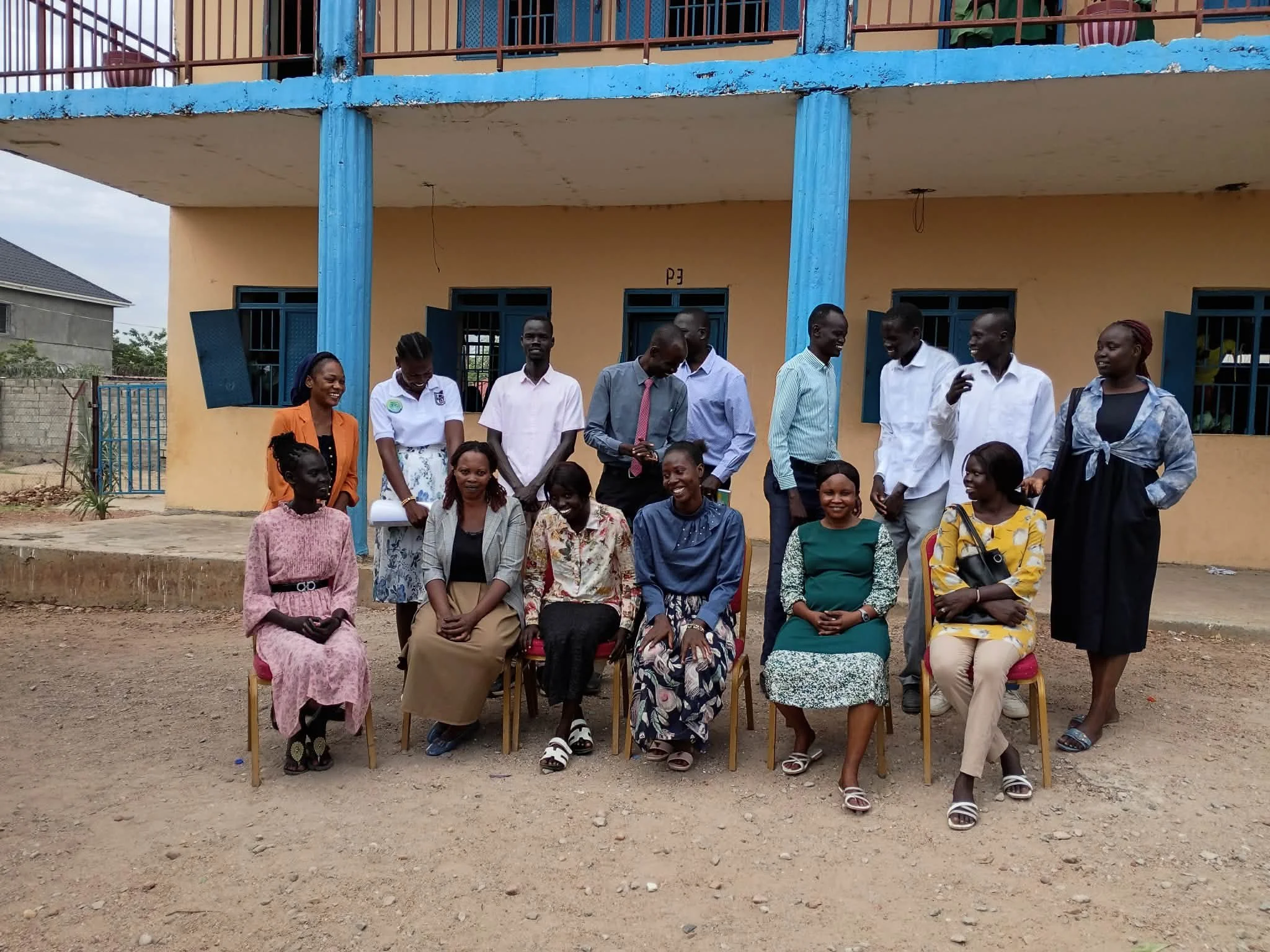 A group of women and men, some seated and some standing, gathered outside a yellow building with blue pillars and windows, smiling and appearing to be part of a community event.