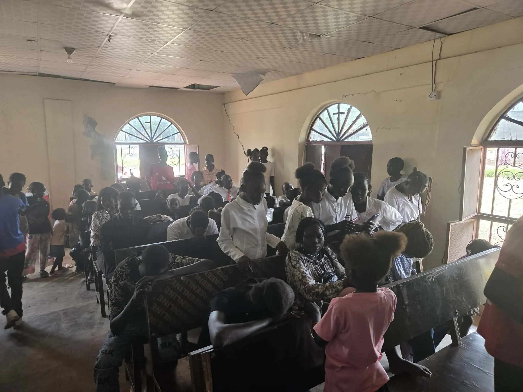 A group of people gathered inside a room with arched windows, some seated and some standing, during daytime.