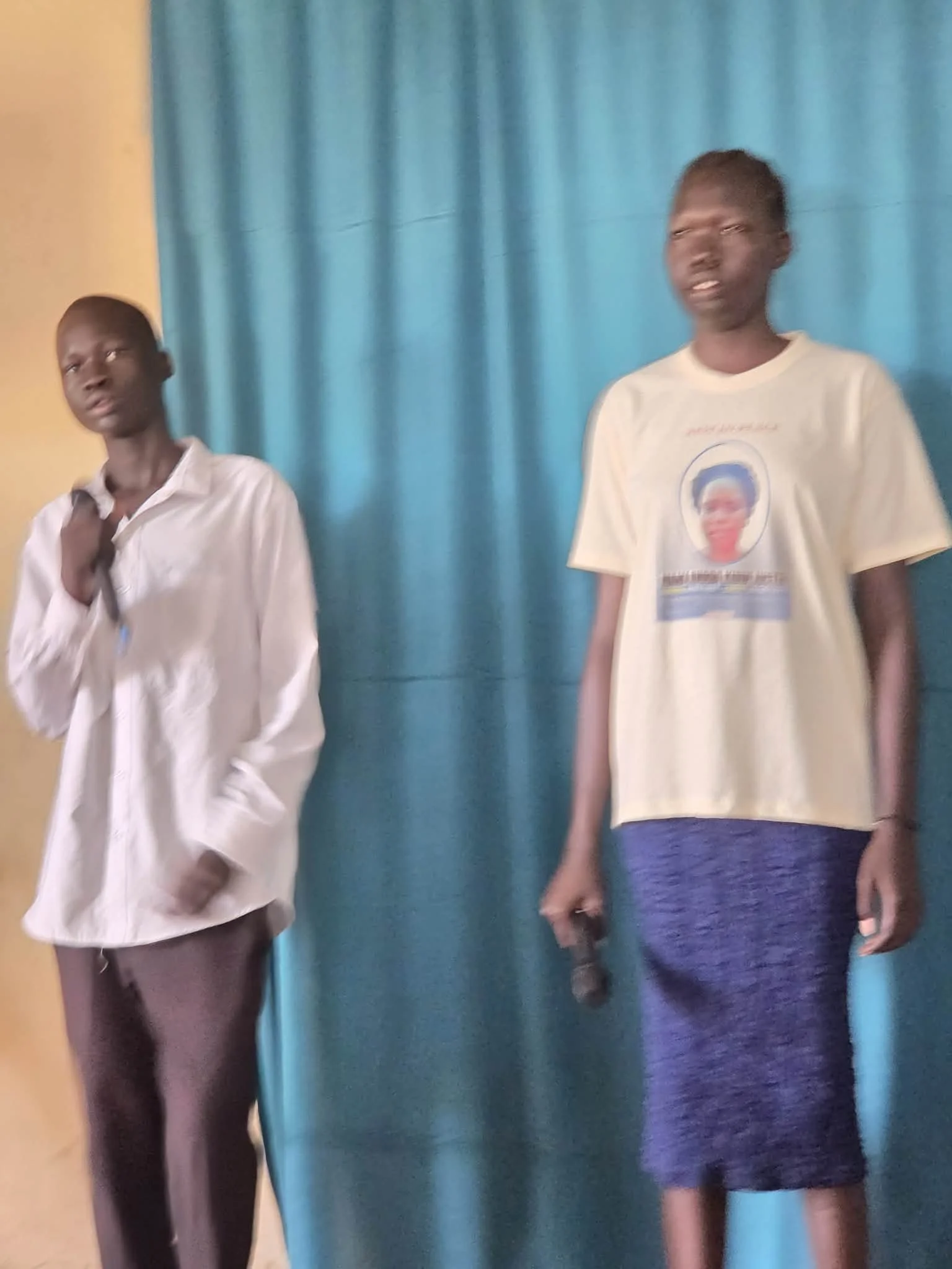 Two boys stand in front of a blue curtain. The boy on the left wears a white shirt and brown pants, and the boy on the right wears a T-shirt with a political campaign image and blue shorts.