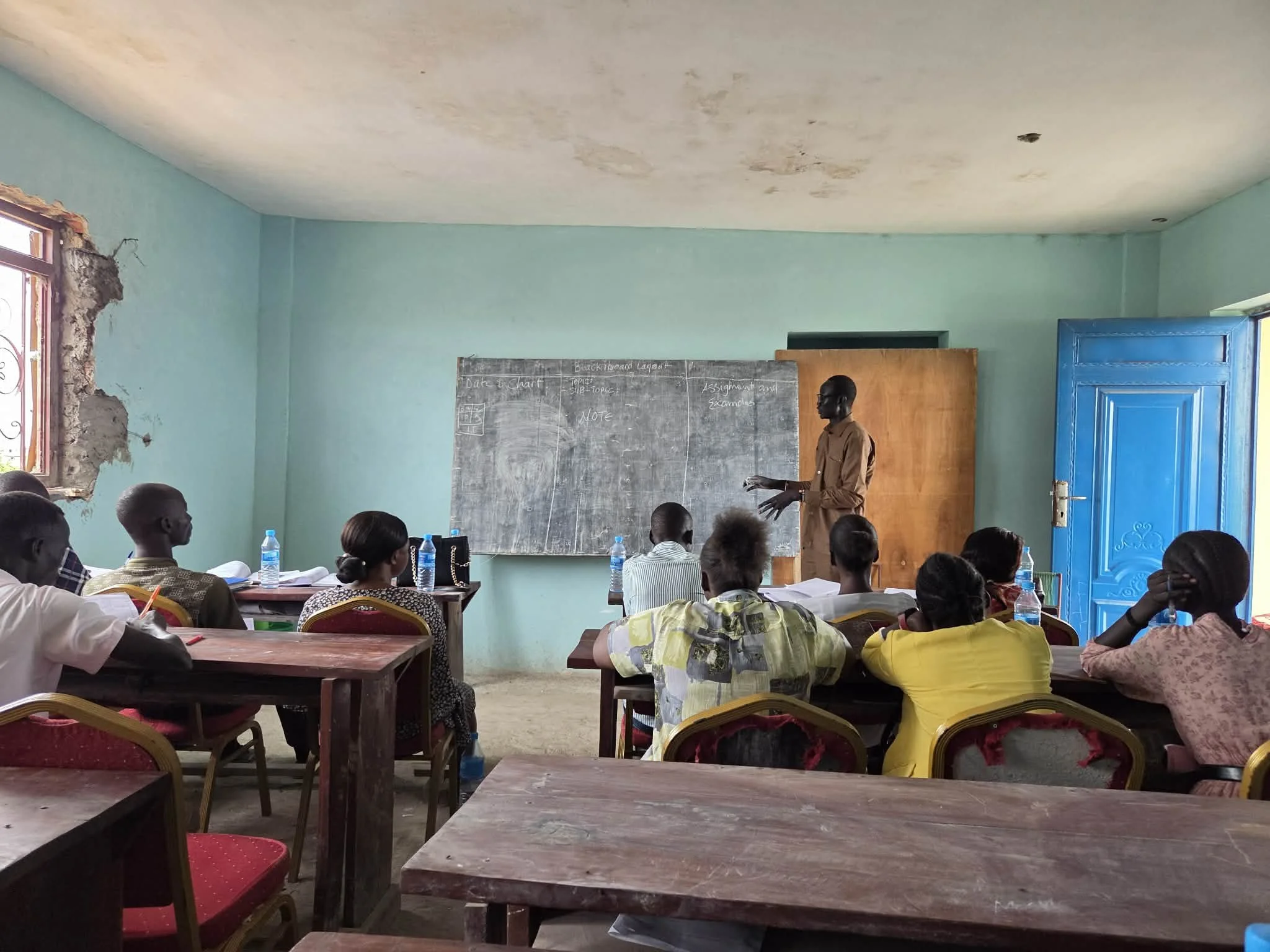 A classroom with students seated at desks, listening to a teacher who is standing near a chalkboard in front of the room. The room has blue walls and a partially broken window, with some water bottles on the desks.