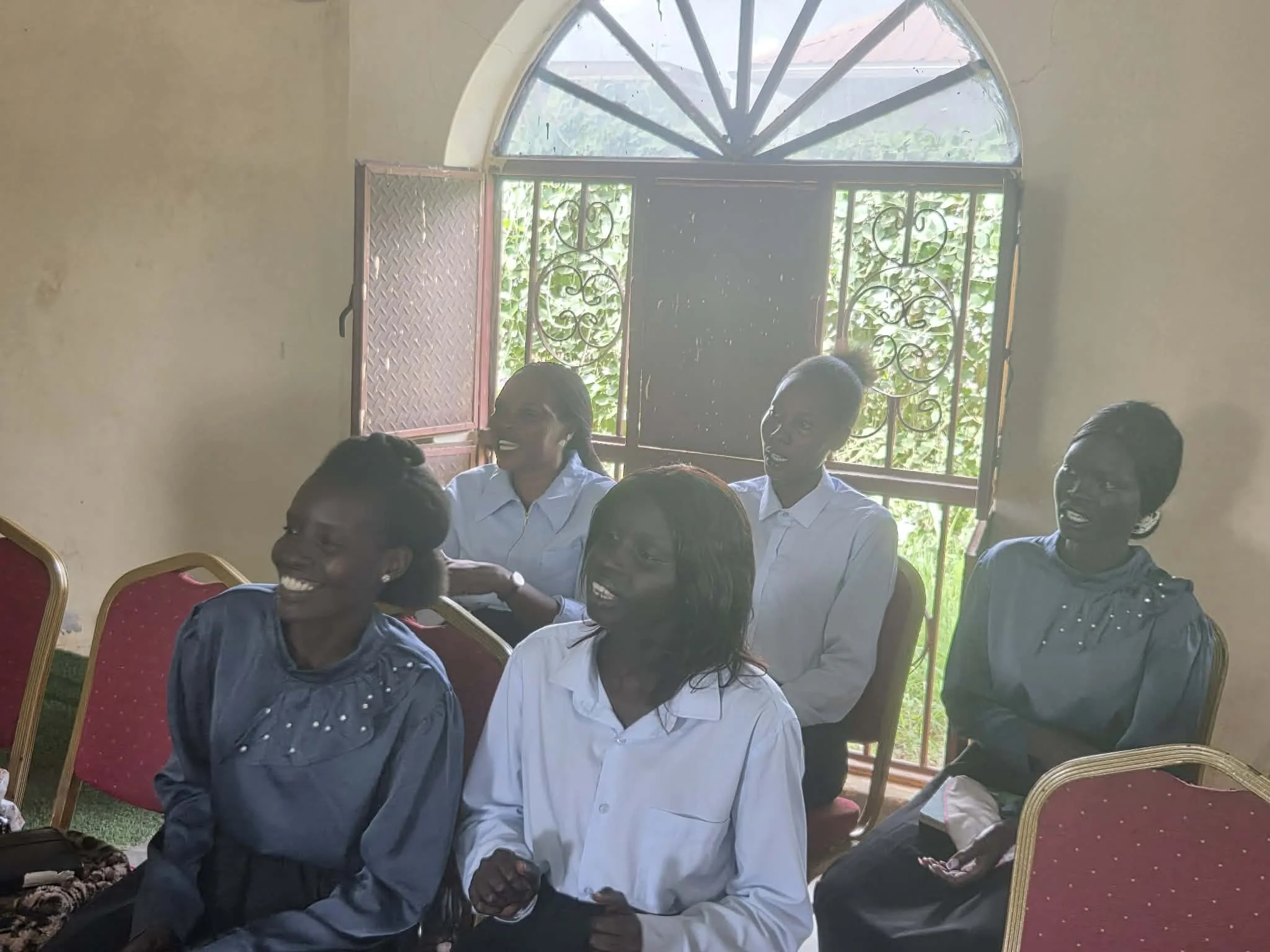 A group of smiling people wearing white shirts gathered indoors near a window with decorative iron bars, with some chairs visible around them.