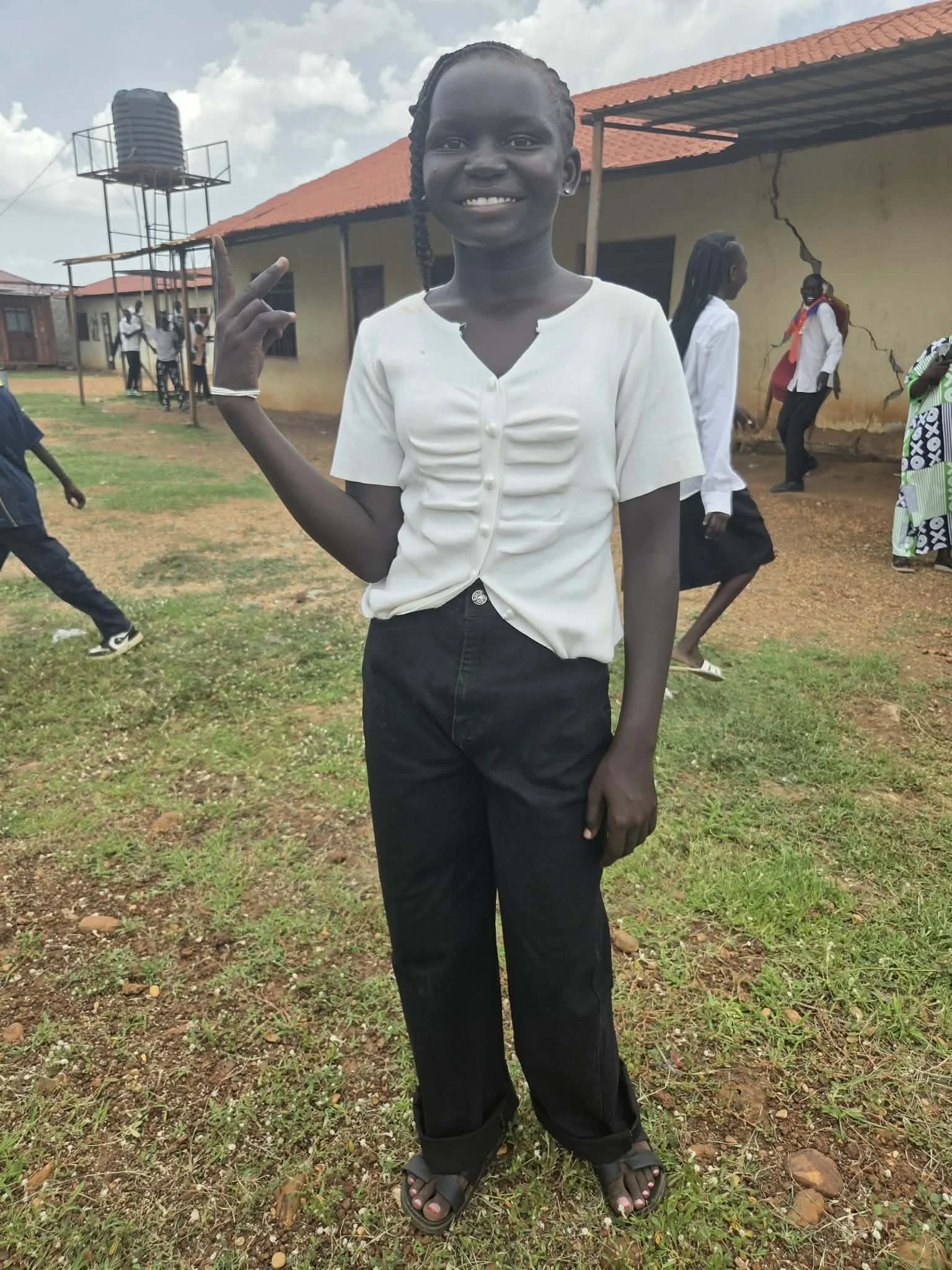 A smiling young girl with braided hair wearing a white button-up shirt and black pants standing outdoors on a patchy grassy ground with other children and a building with a cracked wall in the background.