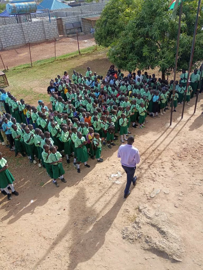 School children gathered in lines for assembly under a tree, with a teacher or supervisor supervising, and a man walking towards them on a dirt ground.