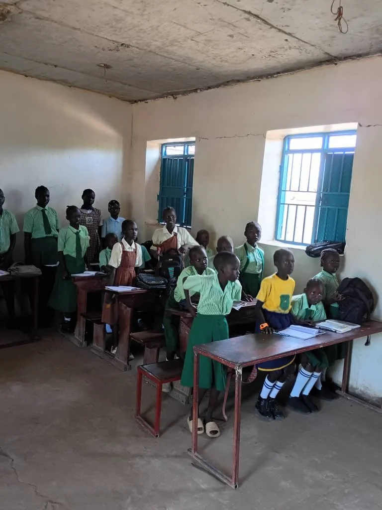 A classroom with students in uniforms, some standing and some sitting at desks, inside a room with unpainted walls and two windows.
