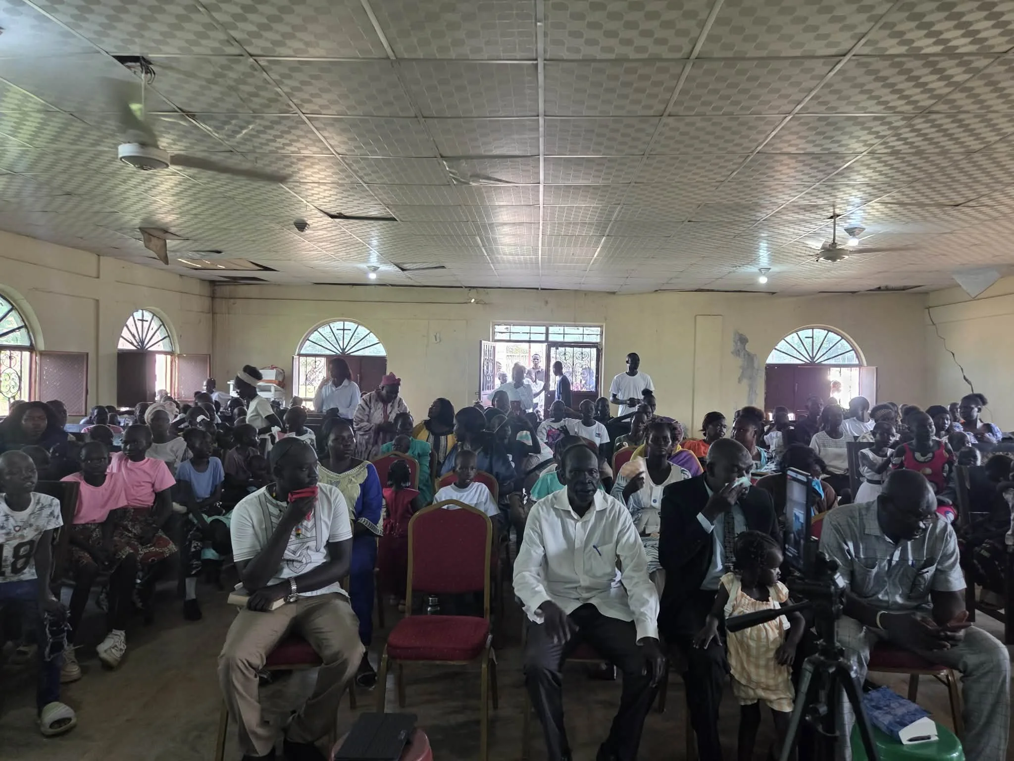 Crowd of people gathered inside a community hall with high ceilings, ceiling fans, and large arched windows.