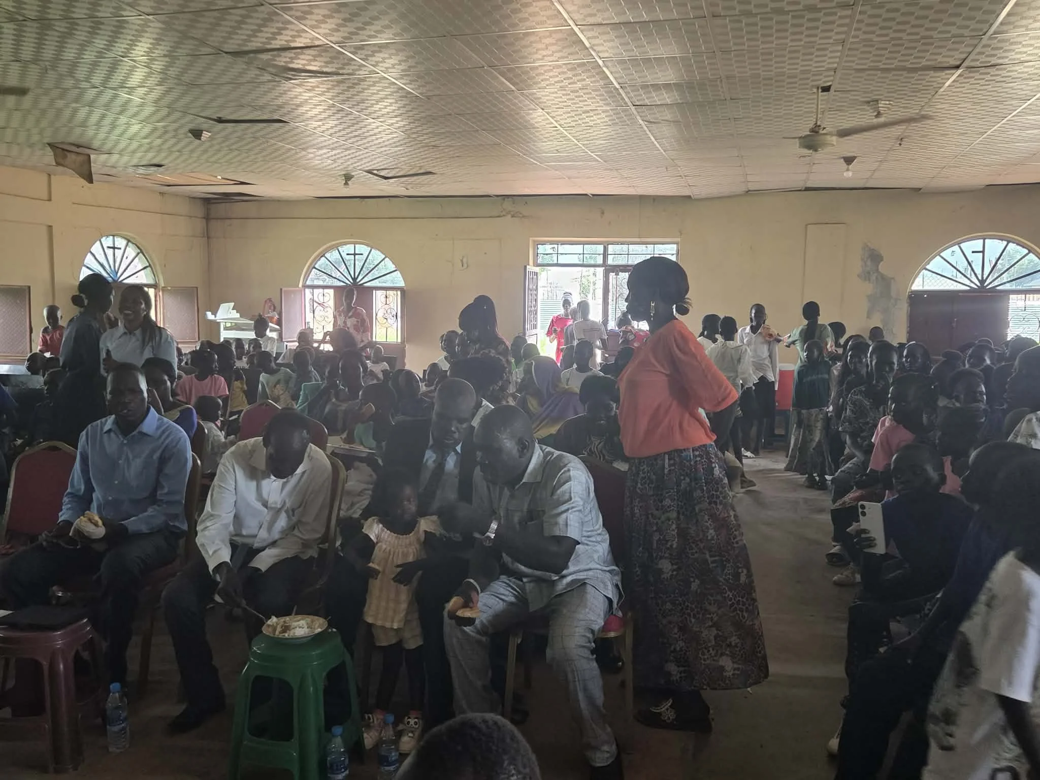 A large room filled with many people sitting and standing, with some people smiling and talking. The room has arched windows, a high ceiling with ceiling fans, and a few women and children are visible. Some individuals are eating, and the overall sce