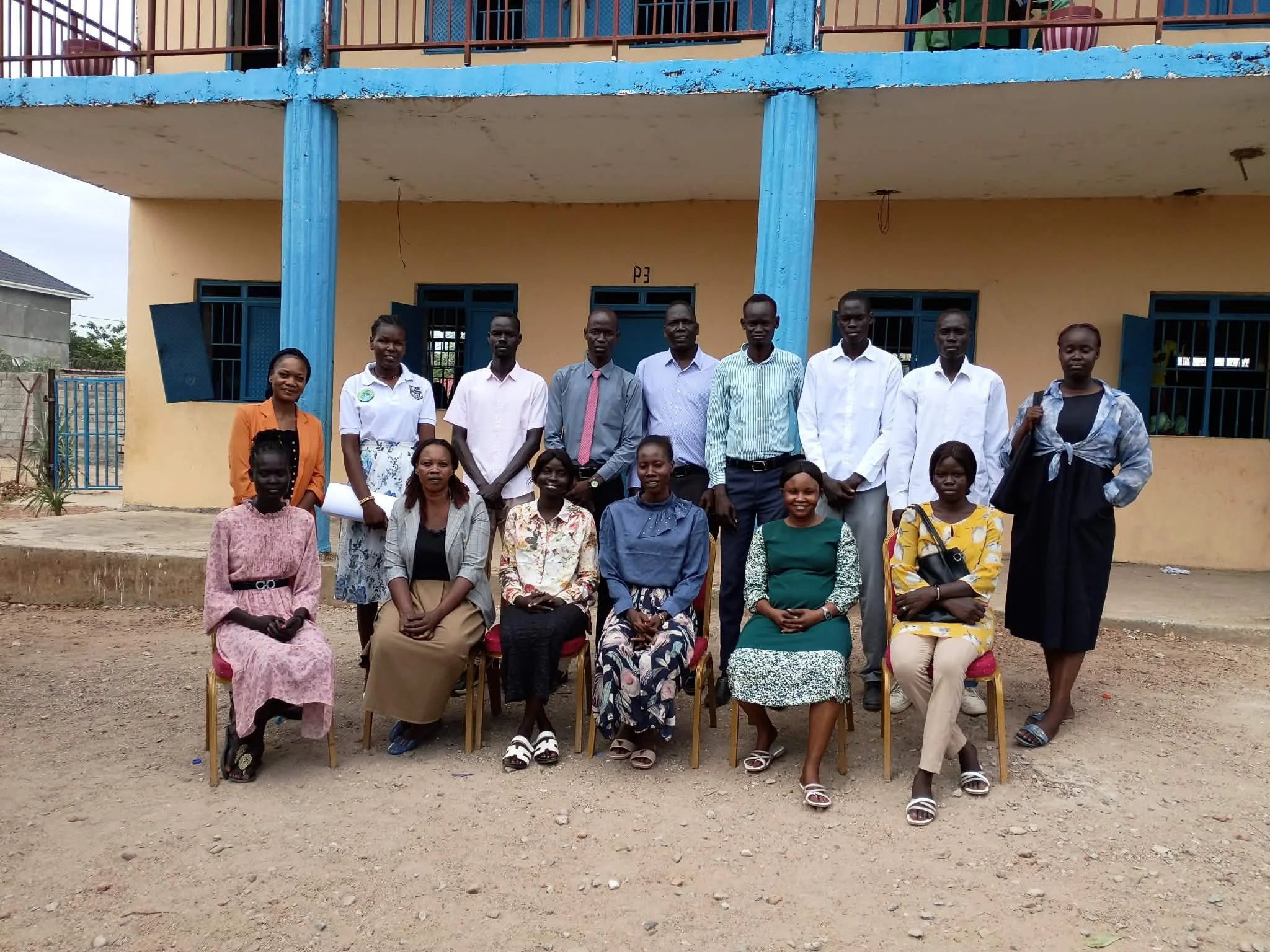 Group of people, including children and adults, posing for a photo in front of a building with yellow walls and blue pillars. The group is standing and sitting on chairs, outdoors on a dirt ground.