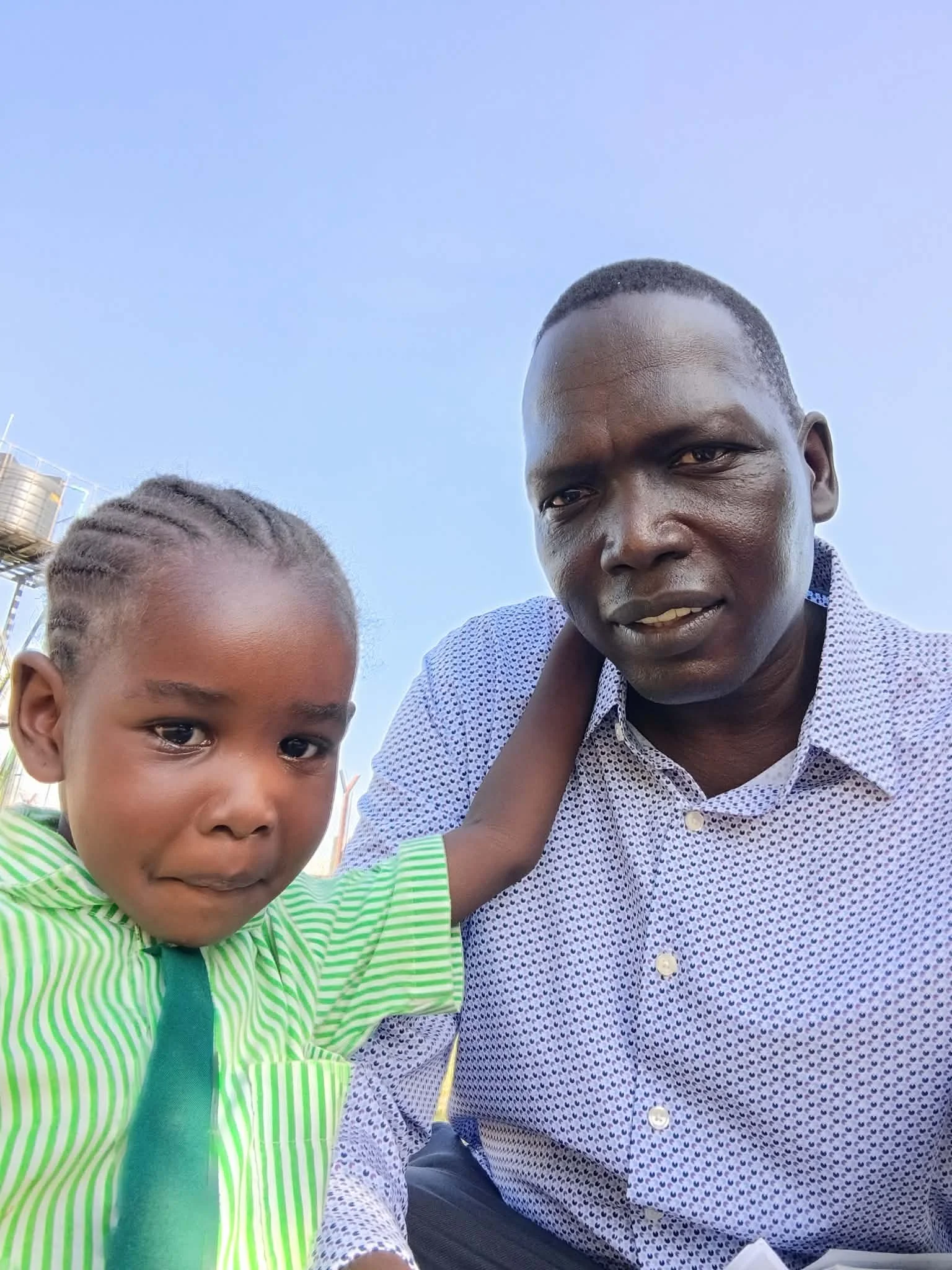 A man and a young boy are taking a selfie together outdoors against a clear blue sky. The man is wearing a patterned shirt, and the boy is wearing a green and white striped shirt with a dark green strap.