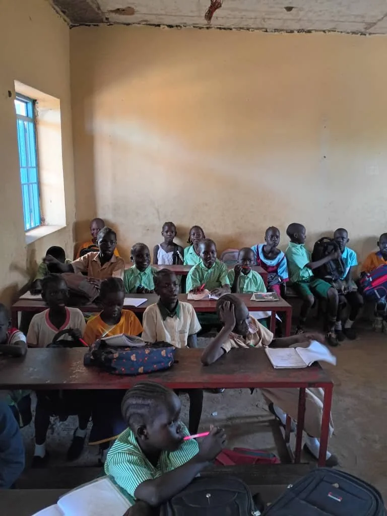 A classroom with students sitting at desks, some with books and notebooks, inside a simple room with a window and a partially unfinished ceiling.