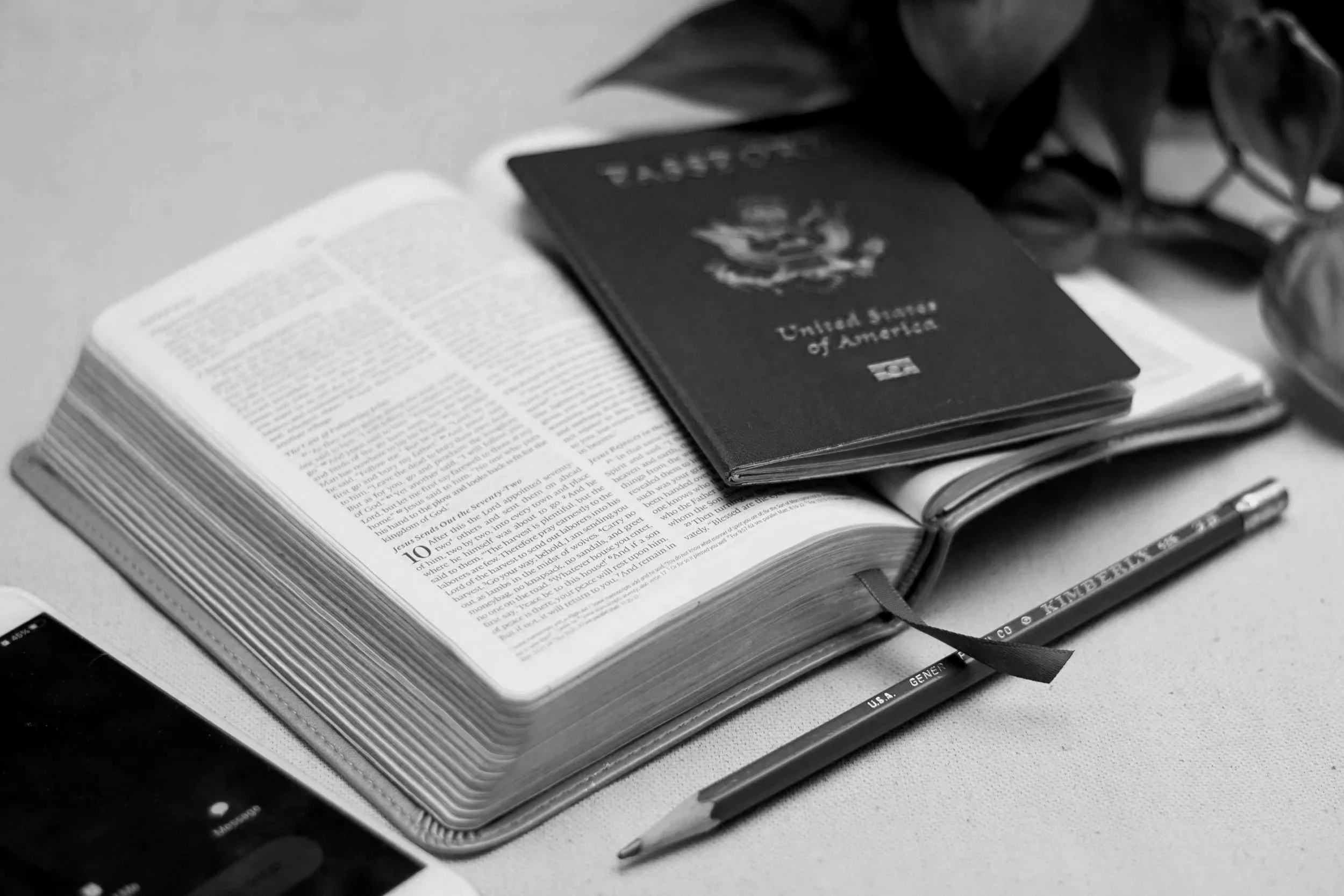 An open Bible, a United States passport resting on top of it, a pencil, and some plants on a table.