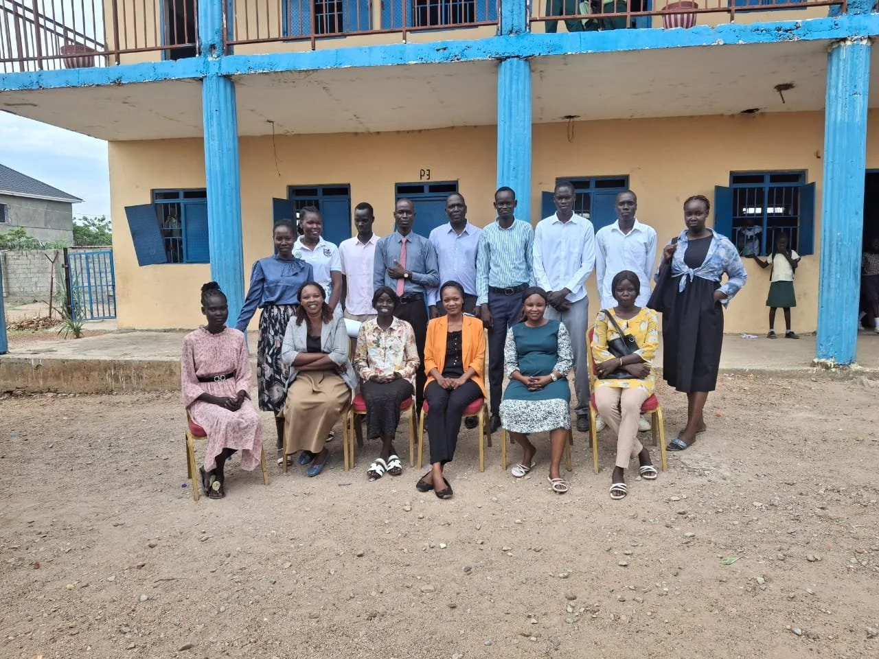 Group of people, men and women, posing for a photo in front of a building with a yellow wall and blue pillars, on a dirt ground.