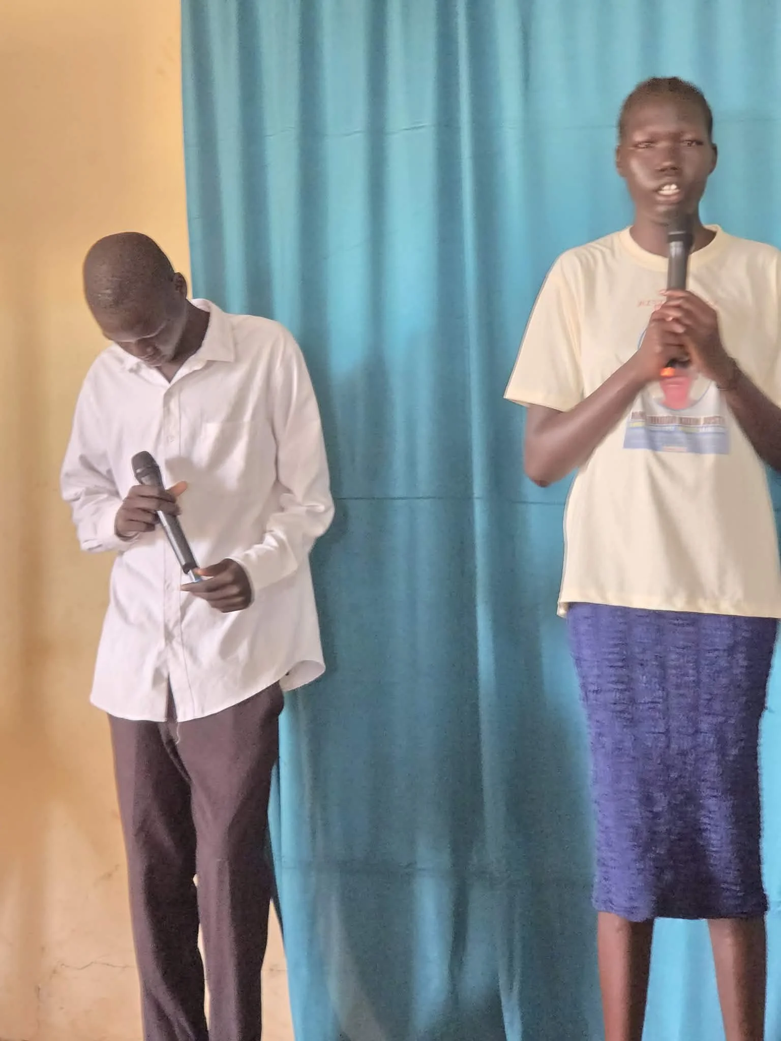 Two boys standing in front of blue curtain, holding microphones. One boy is wearing a white shirt and dark pants, looking down. The other boy is wearing a light-colored shirt and a blue skirt or shorts, speaking into the microphone.