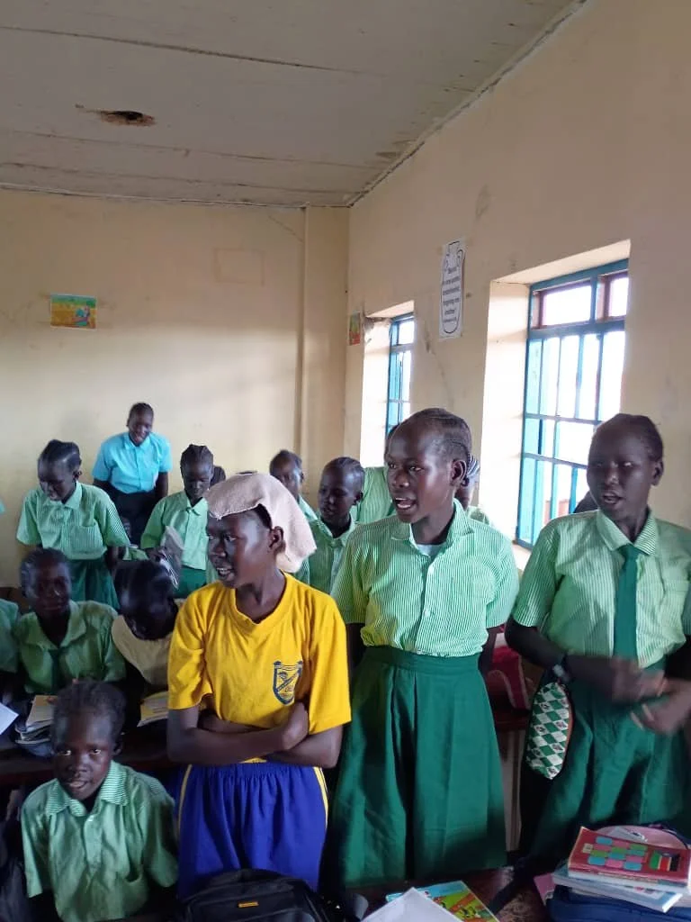 School classroom with students in green uniforms and one girl in yellow, standing and talking, with a teacher in the background. The room has large windows with bars and posters on the walls.