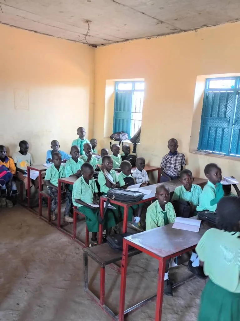 Children in green school uniforms sitting at desks in a classroom with yellow walls and blue window shutters.