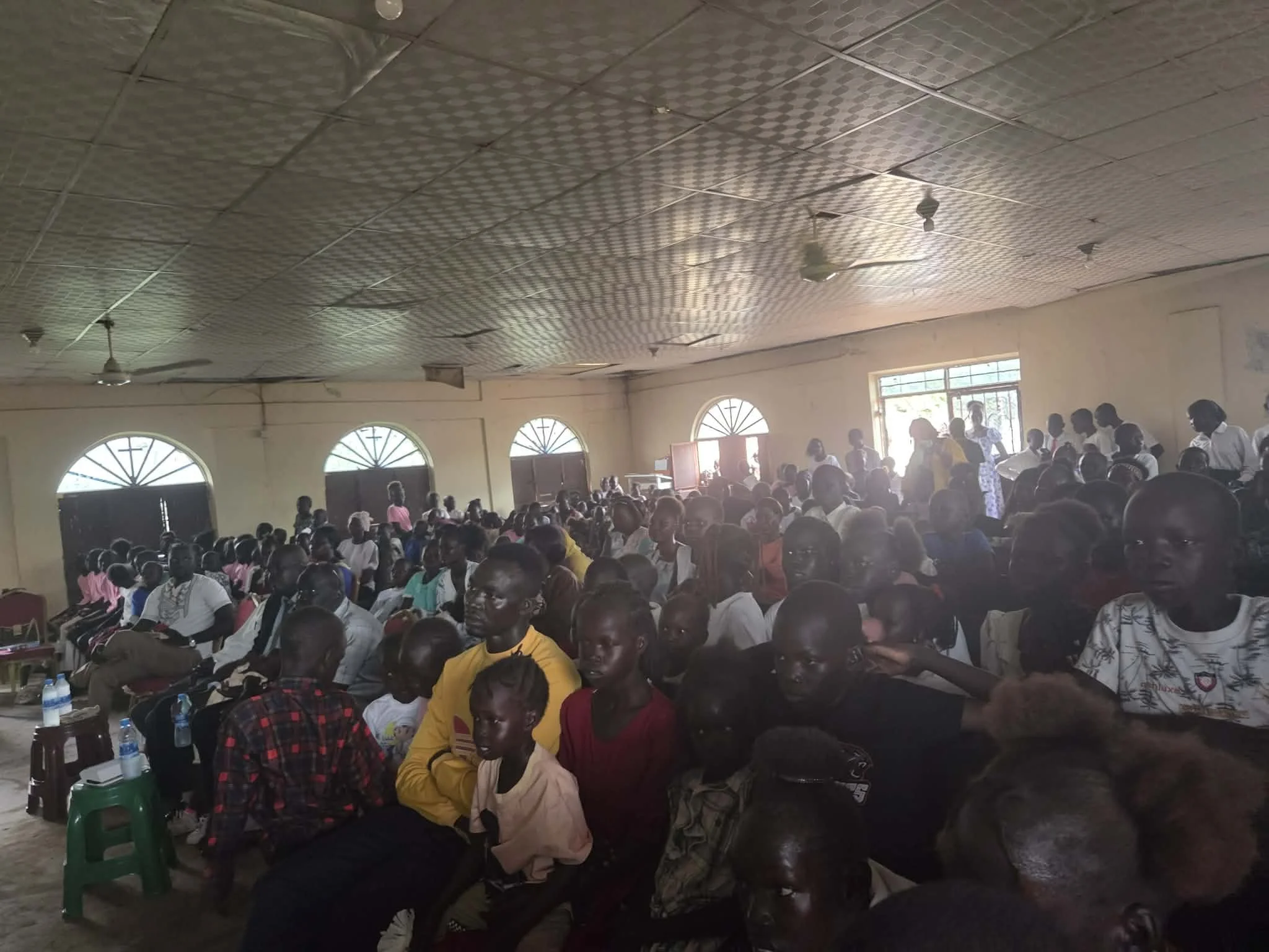 A large group of people sitting in a hall attending an event. The hall has a decorated ceiling with ceiling fans, and windows allowing natural light. The crowd includes men, women, and children.