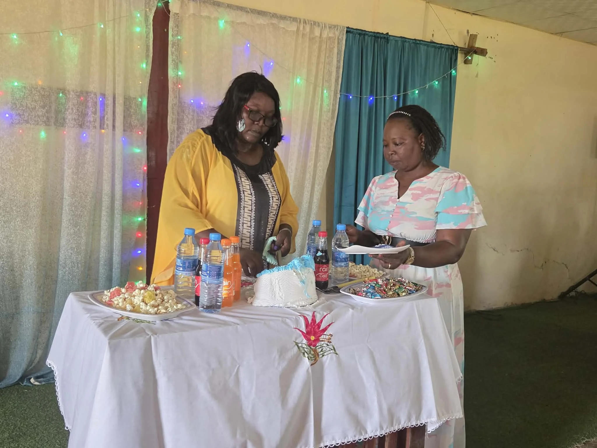 Two women standing behind a table with snacks and drinks, with string lights and curtains in the background.
