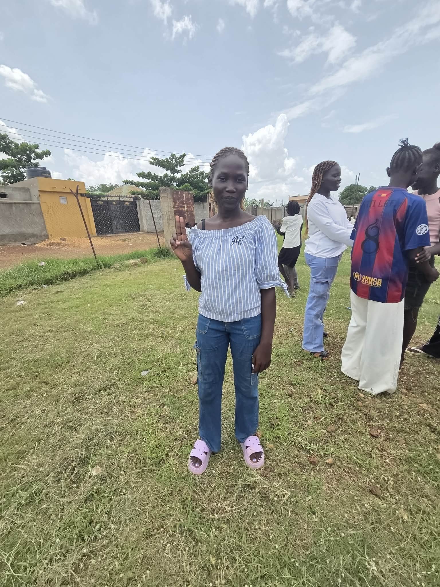 A woman standing outdoors on grass, making a peace sign with her right hand, wearing a blue striped top, jeans, and pink sandals, with other people in the background.