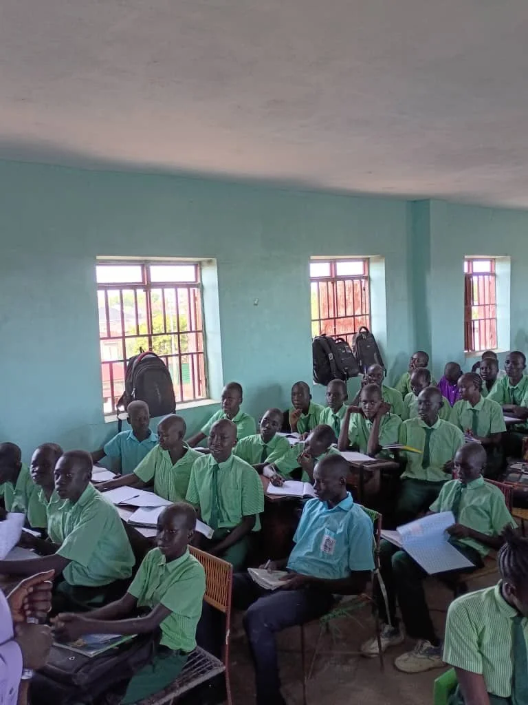 Students sitting at desks in a classroom, wearing green uniforms, with backpacks on windowsills.